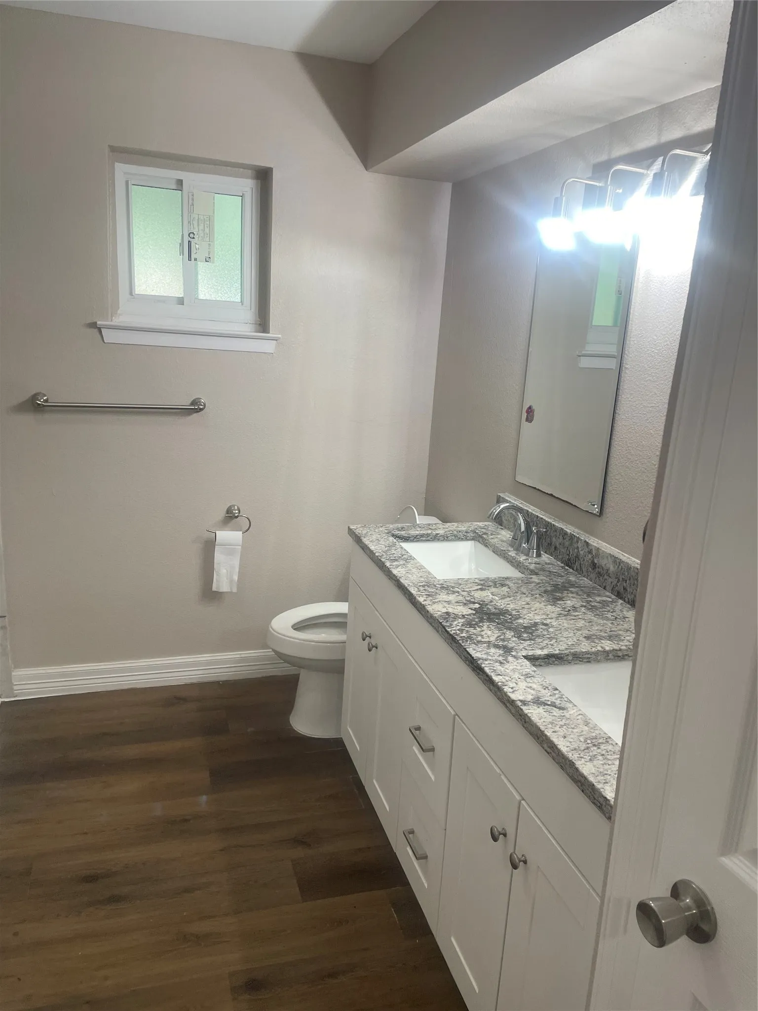 Wood-finish flooring, dual basin vanity with white cabinetry, stone countertop, window, and wall-mounted lighting