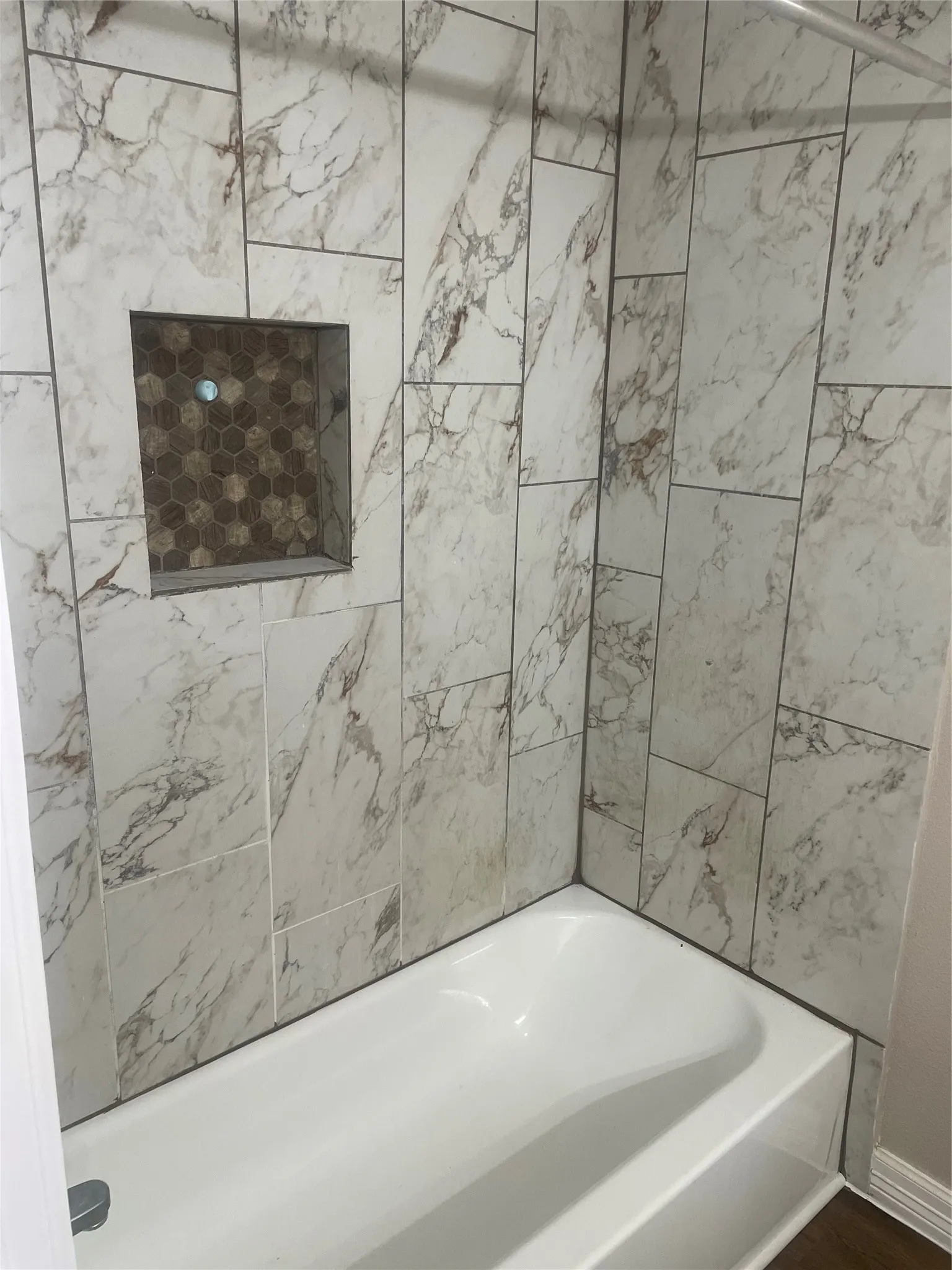 Bathtub with a white finish, white and brown variegated tile surround, and a recessed niche featuring hexagonal mosaic tile
