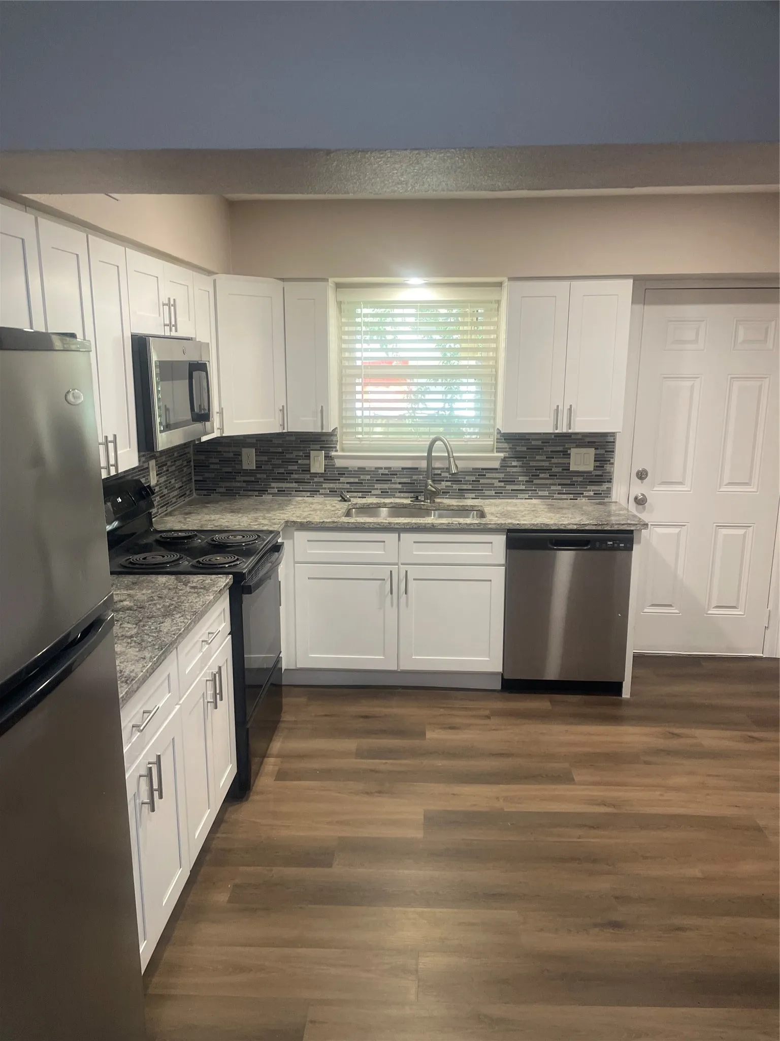 White cabinetry with brushed nickel hardware, stainless steel appliances, mosaic tile backsplash, wood-finish flooring, and light-toned countertops