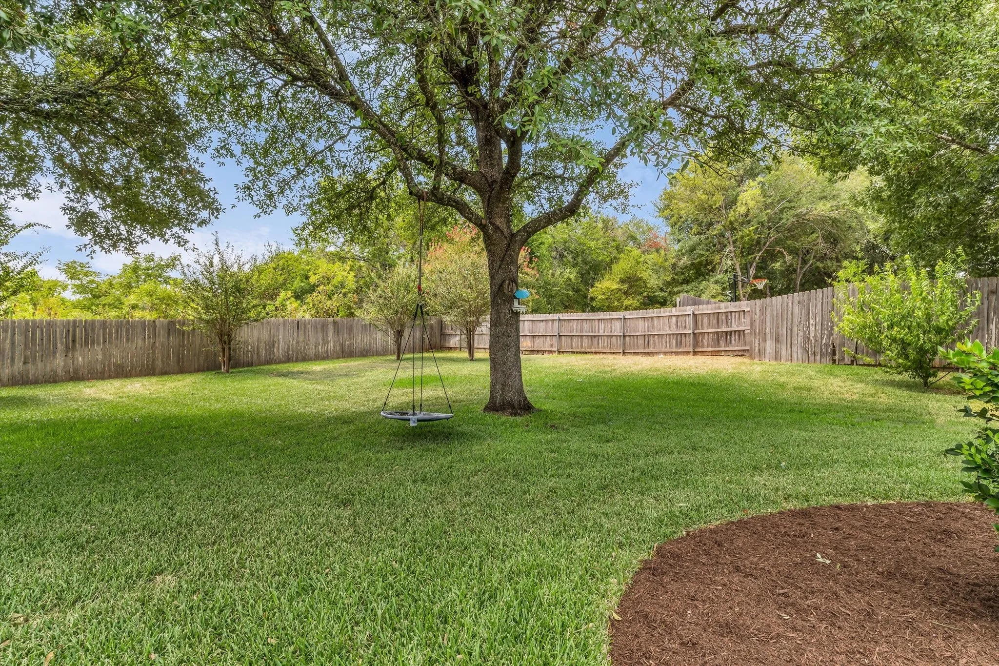 Expansive backyard featuring a mature shade tree, a wooden privacy fence, a manicured lawn, and a mulched planting area