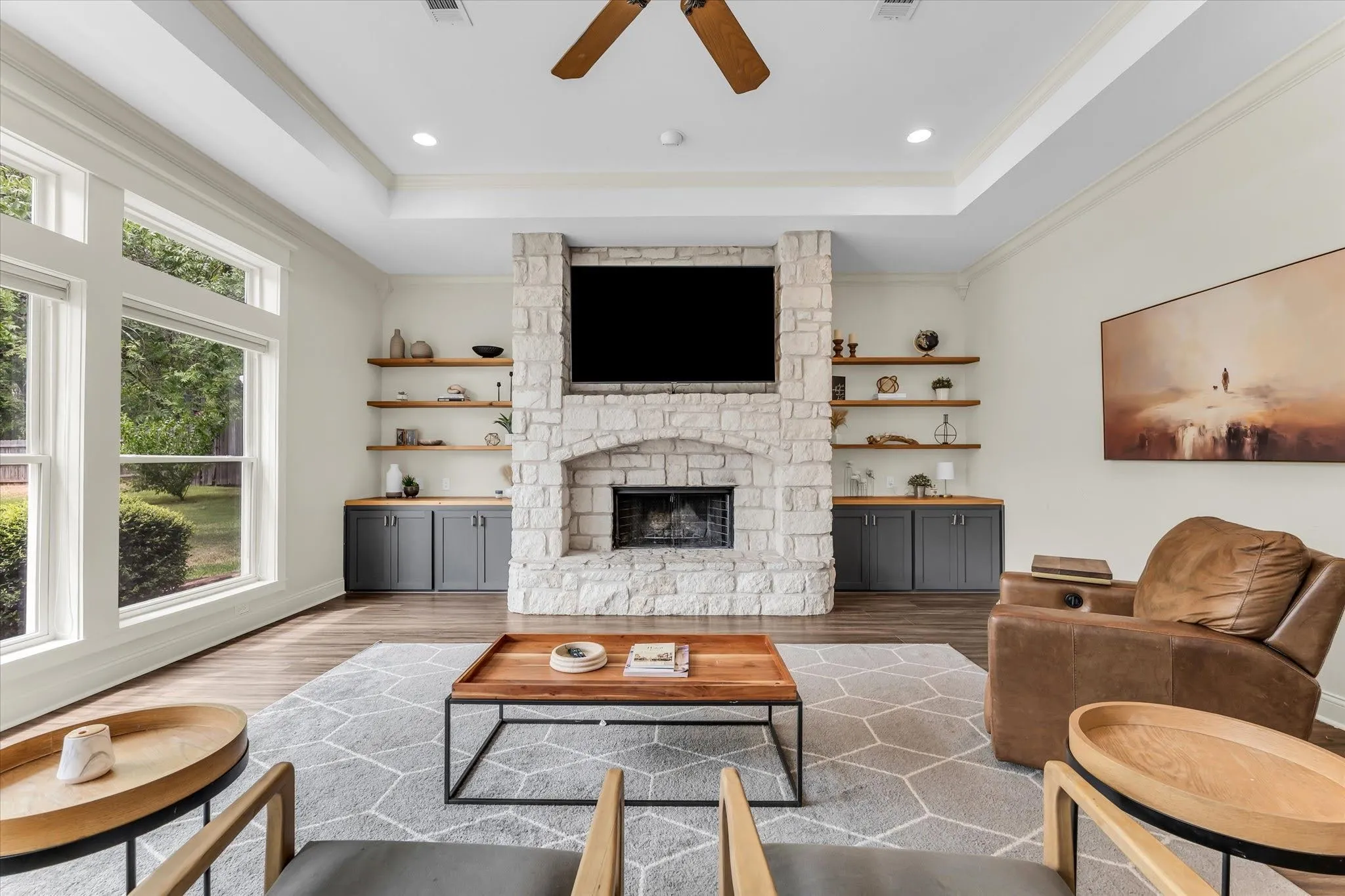 Tray ceiling with recessed lighting and a ceiling fan, stone-faced fireplace, wood-finish flooring, built-in shelving, and built-in cabinetry