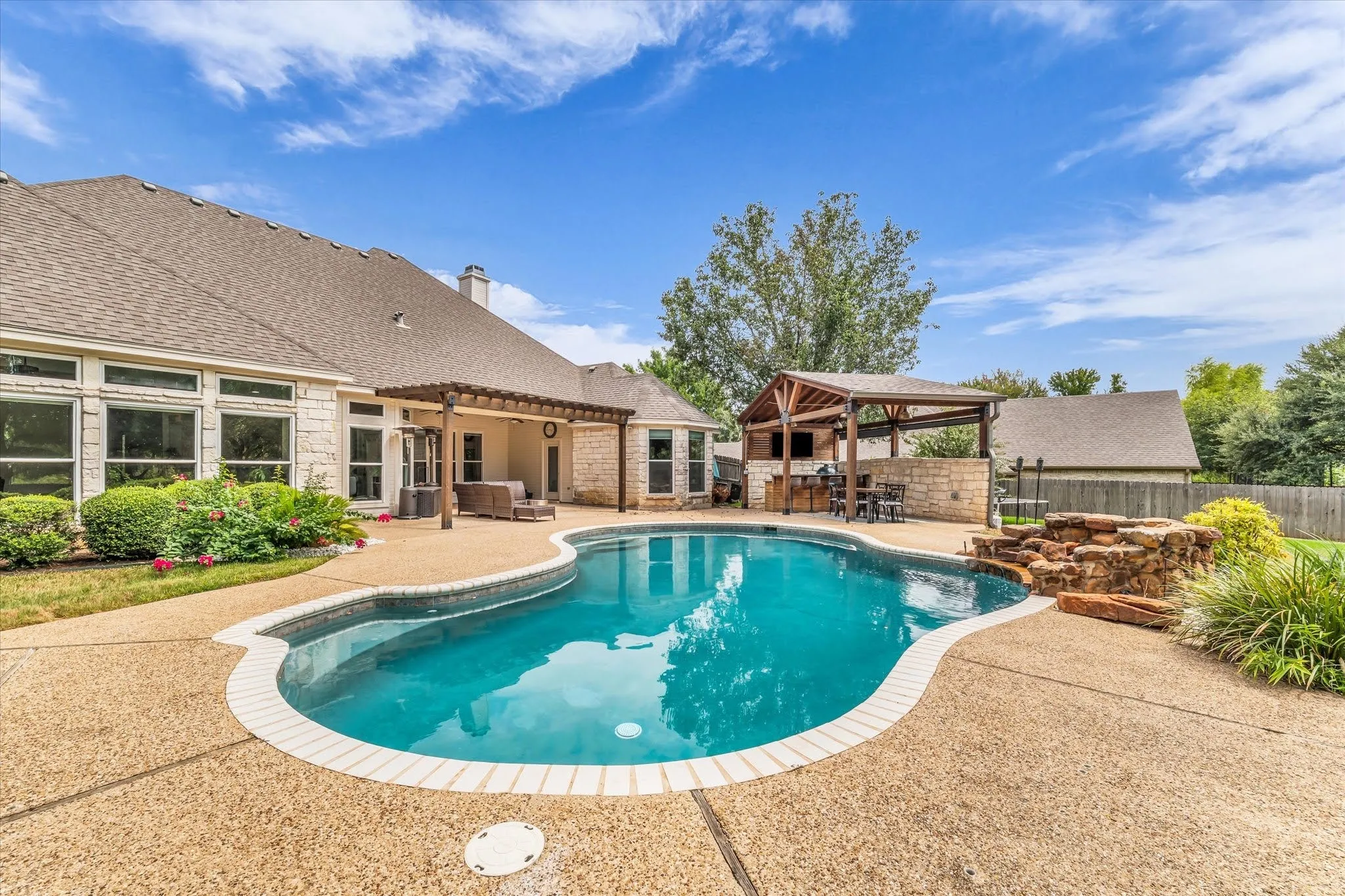 Curved swimming pool with a stone waterfall feature, expansive deck, and a covered outdoor kitchen