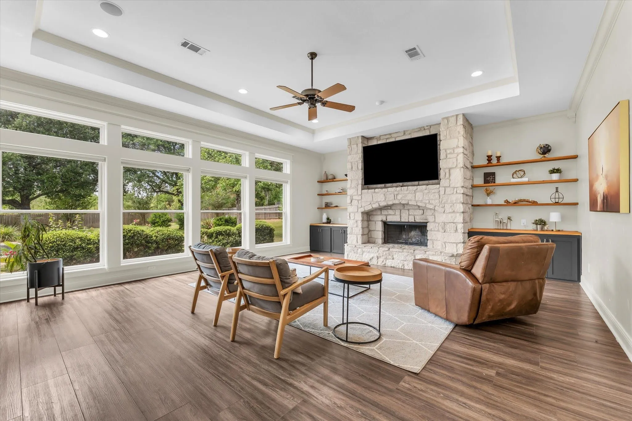Expansive living area featuring a stone-clad fireplace, wood-finish flooring, built-in shelving, a tray ceiling, and large windows