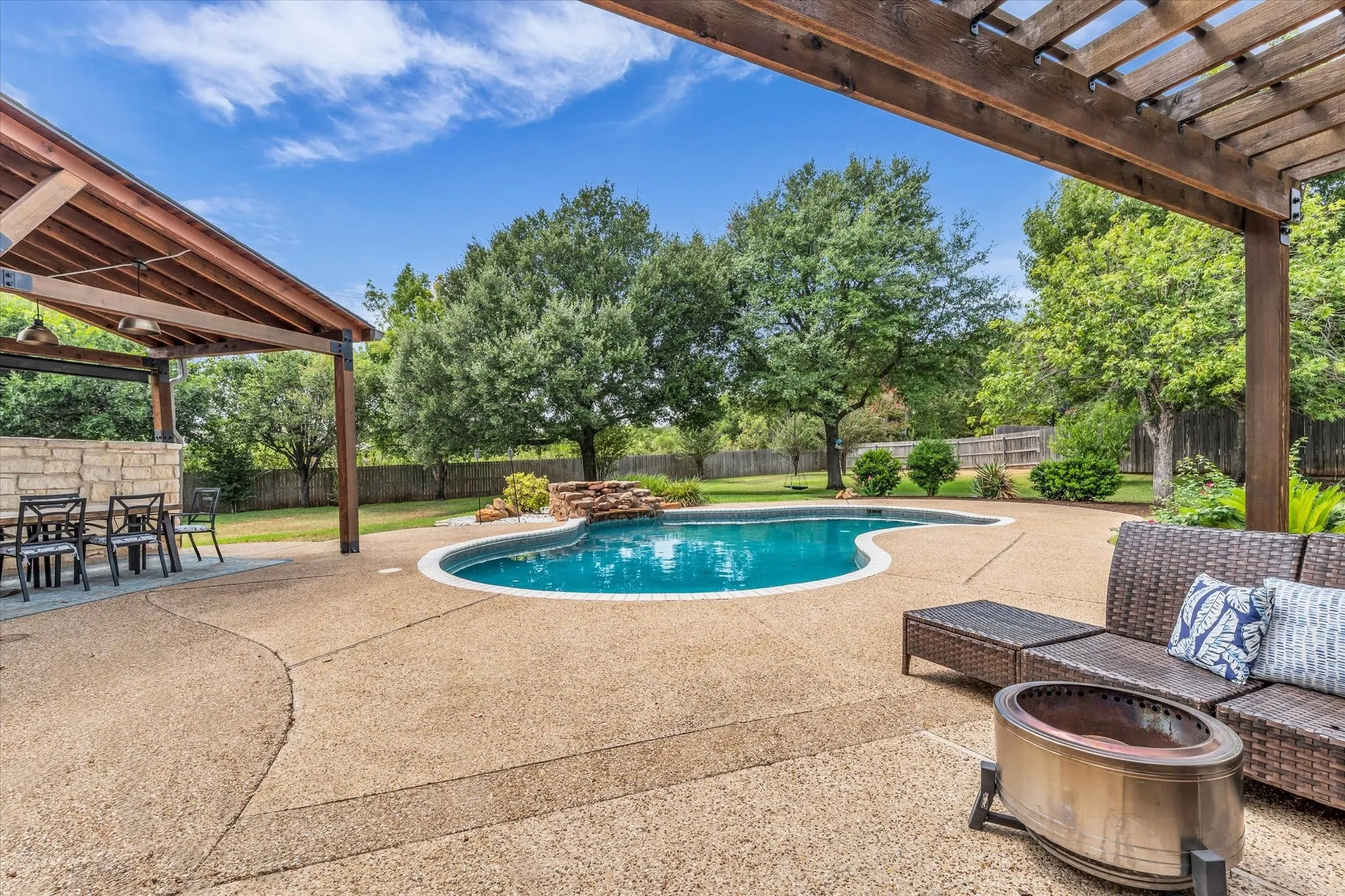Curved swimming pool with a rock water feature, expansive exposed aggregate patio, covered outdoor dining area, and a wood pergola