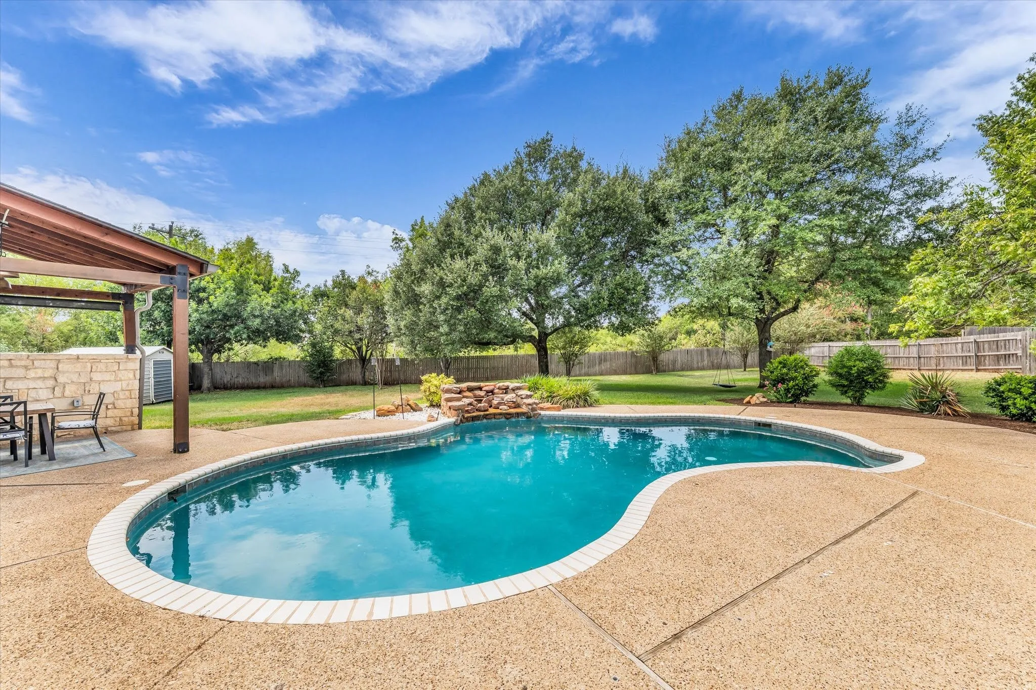 Freeform swimming pool with a stone waterfall feature, aggregate concrete decking, and an attached covered patio with a wood ceiling