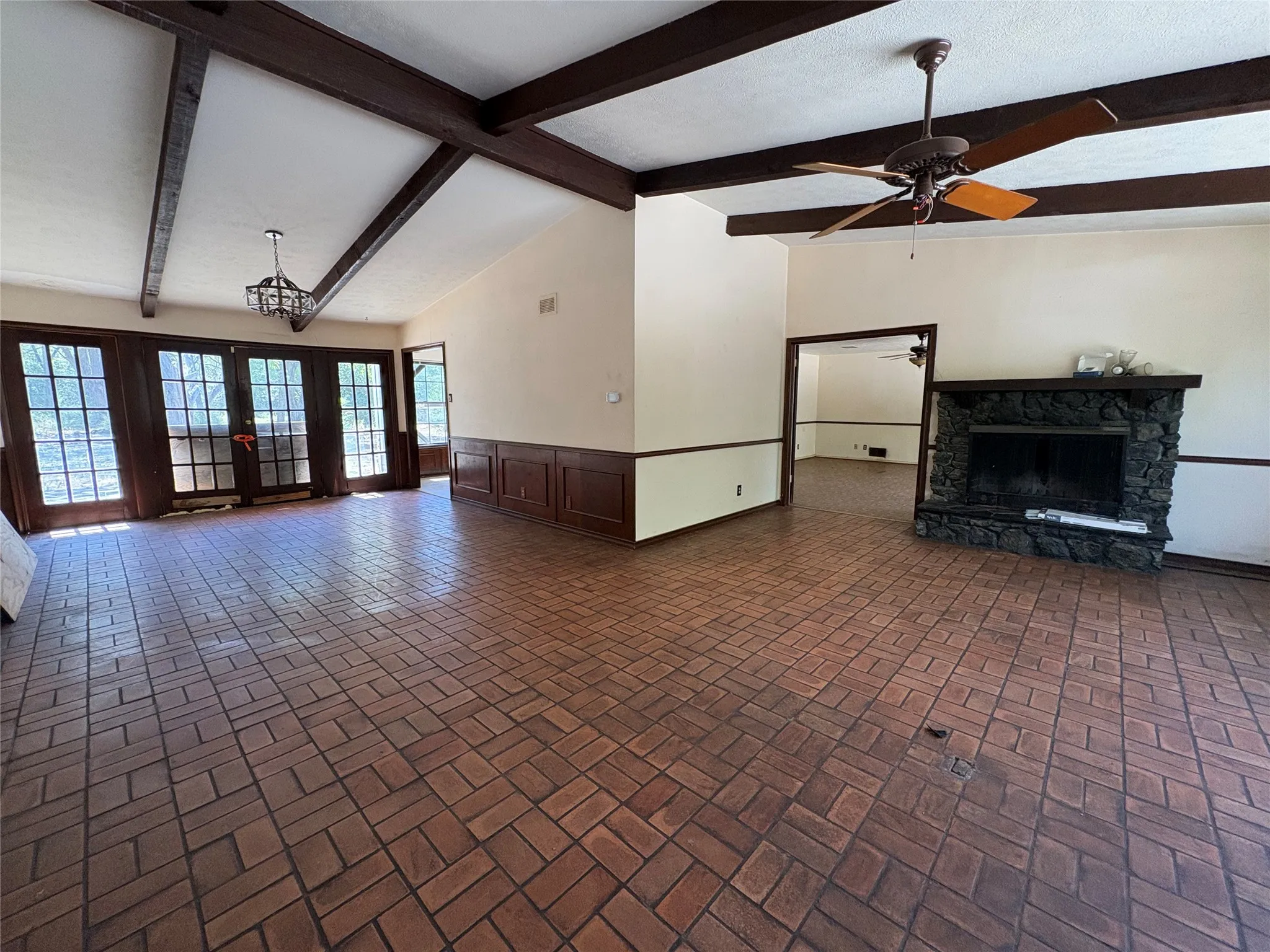 Vaulted ceiling with exposed wood beams, brick-patterned flooring, wood wainscoting, stone-clad fireplace, and multiple sets of French doors