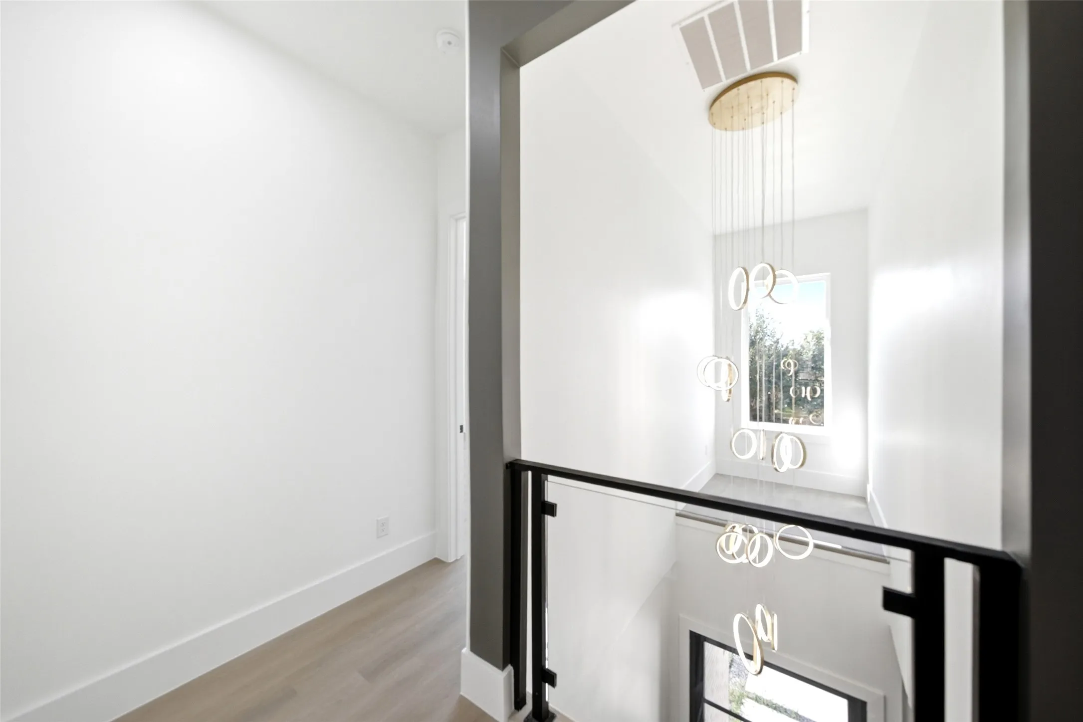 Modern glass panel railing with a dark frame, light wood-finish flooring, and a contemporary multi-ring chandelier