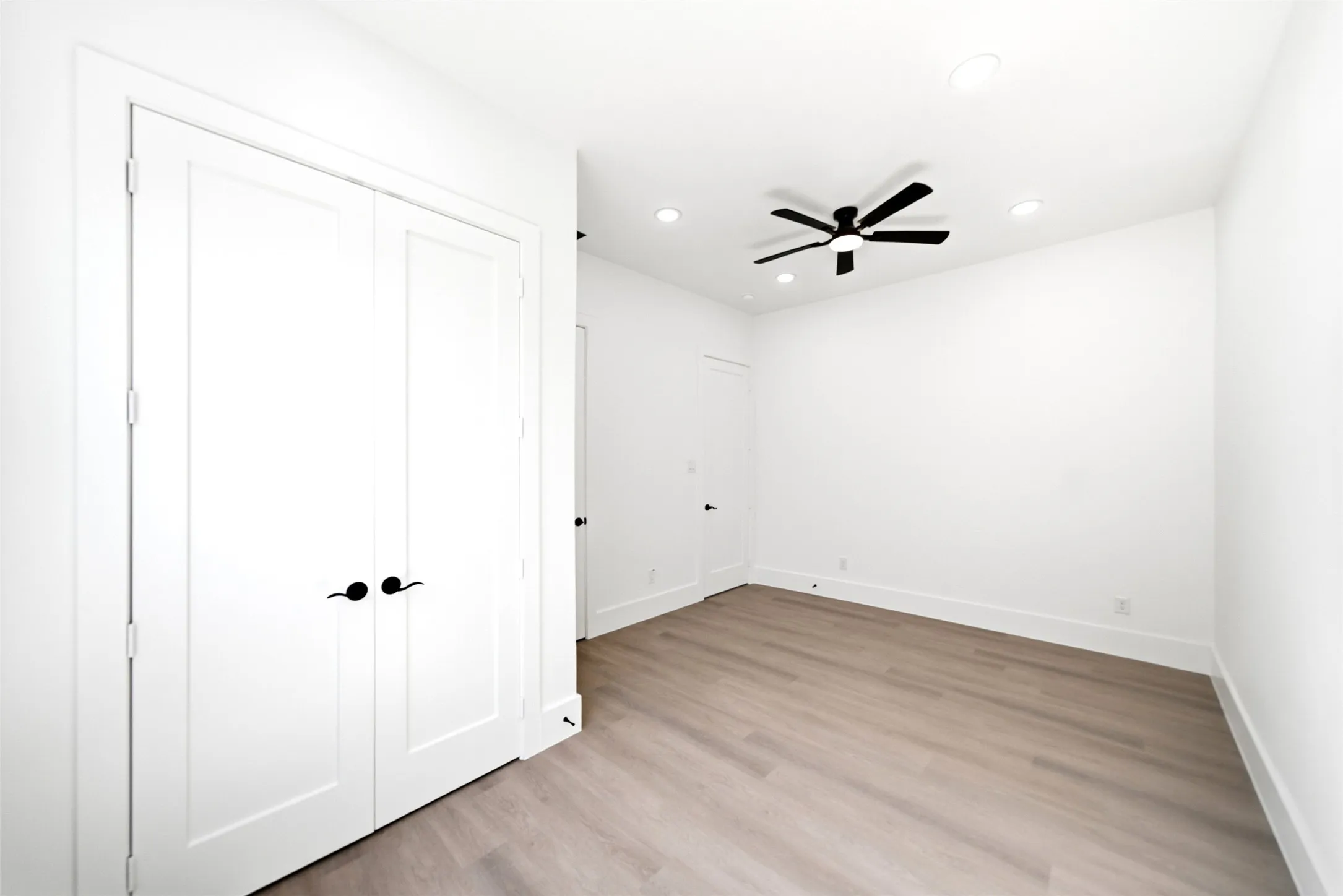 Light wood-finish flooring, white walls, recessed ceiling lighting, and a modern black ceiling fan