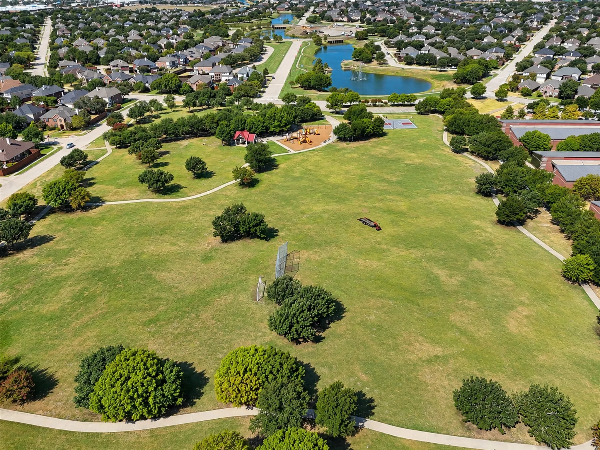 Expansive green space featuring meandering paved pathways, mature trees, a playground area, and a pond