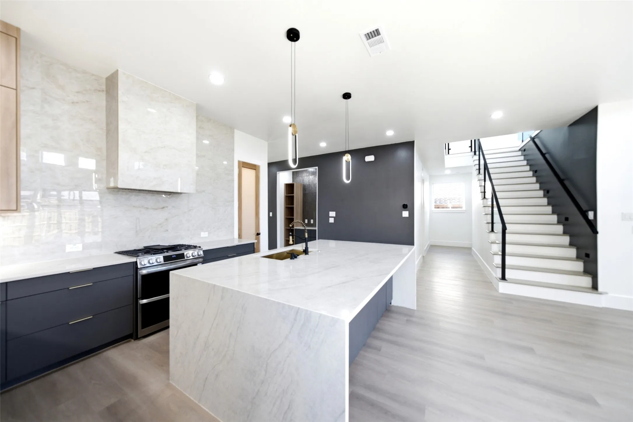 Sleek kitchen featuring an oversized island with waterfall edges, dark flat-panel cabinetry, a gas range with a substantial range hood, and light wood-finish flooring