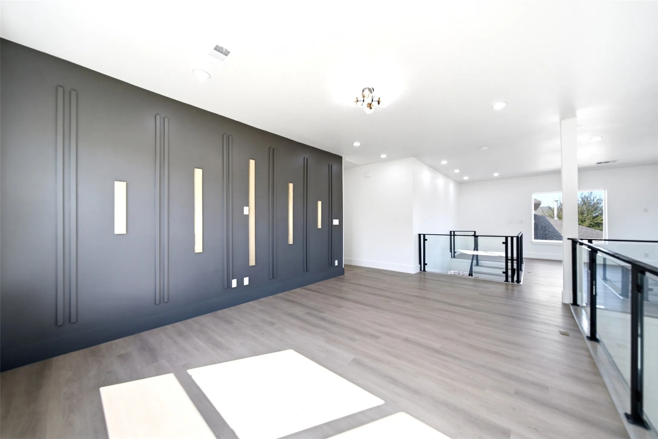 Expansive open-concept space featuring sleek charcoal gray built-in cabinetry with integrated lighting, light wood-finish flooring throughout, recessed ceiling lighting, and floor-to-ceiling windows framing natural light