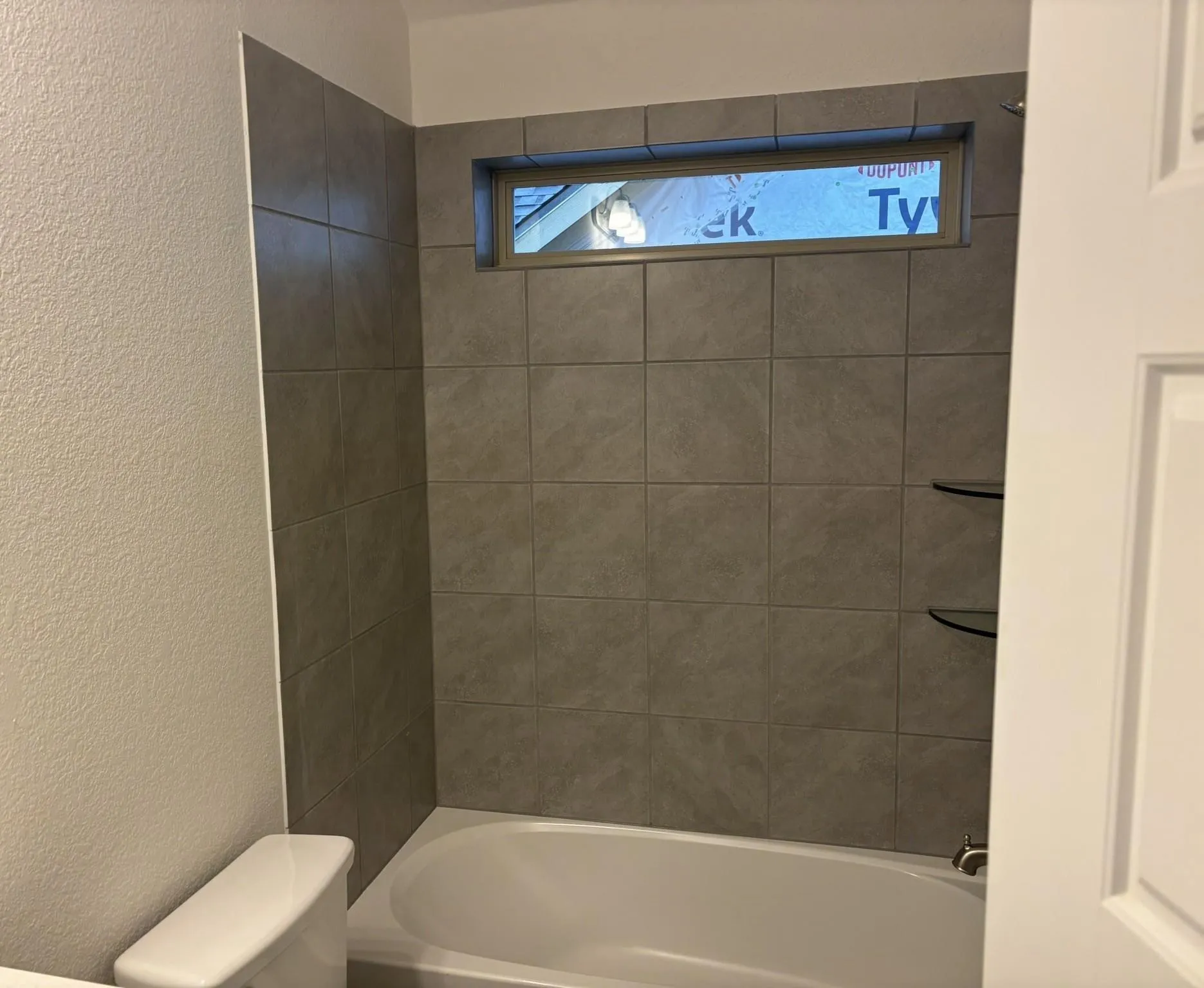 Bathtub with gray tile surround, transom window, and built-in glass shelving