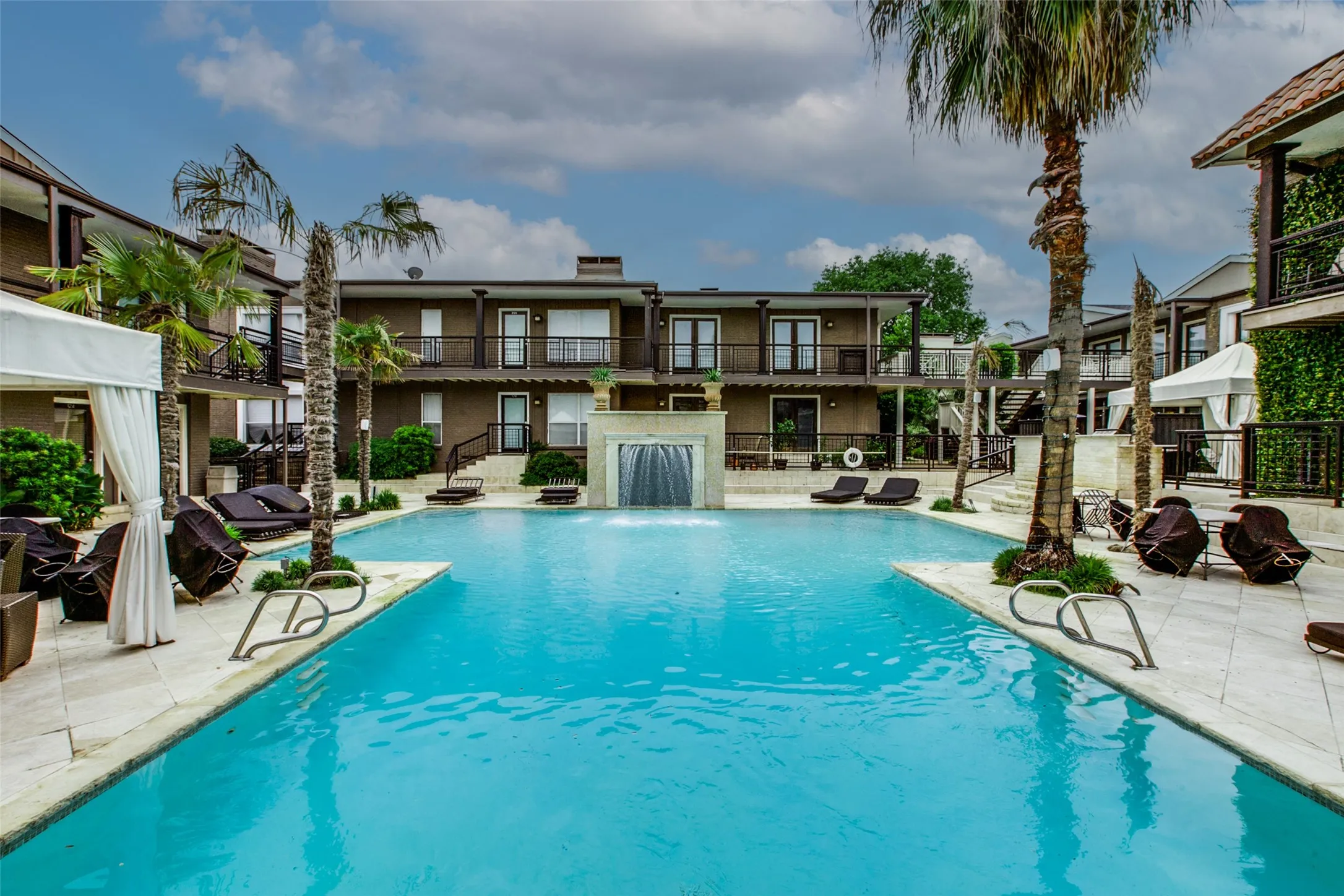 Expansive swimming pool with a central water feature, surrounded by paver decking and mature palm trees