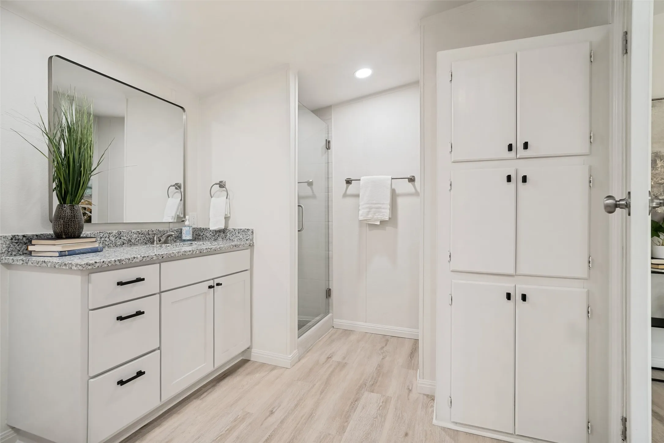 White vanity with a granite countertop, frameless shower enclosure, recessed lighting, and wood-finish flooring