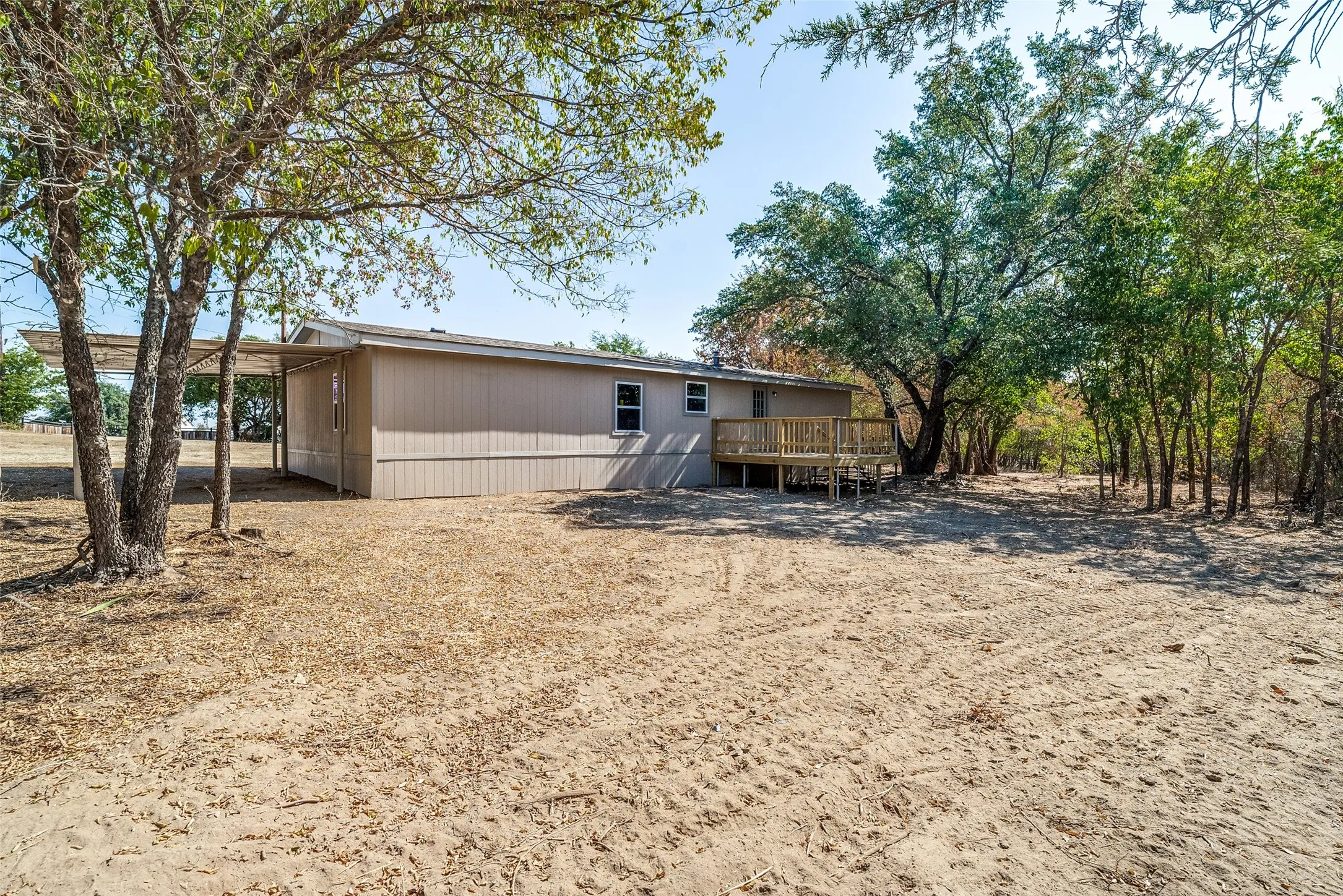 Beige siding exterior with a wood deck, mature trees, and an attached carport