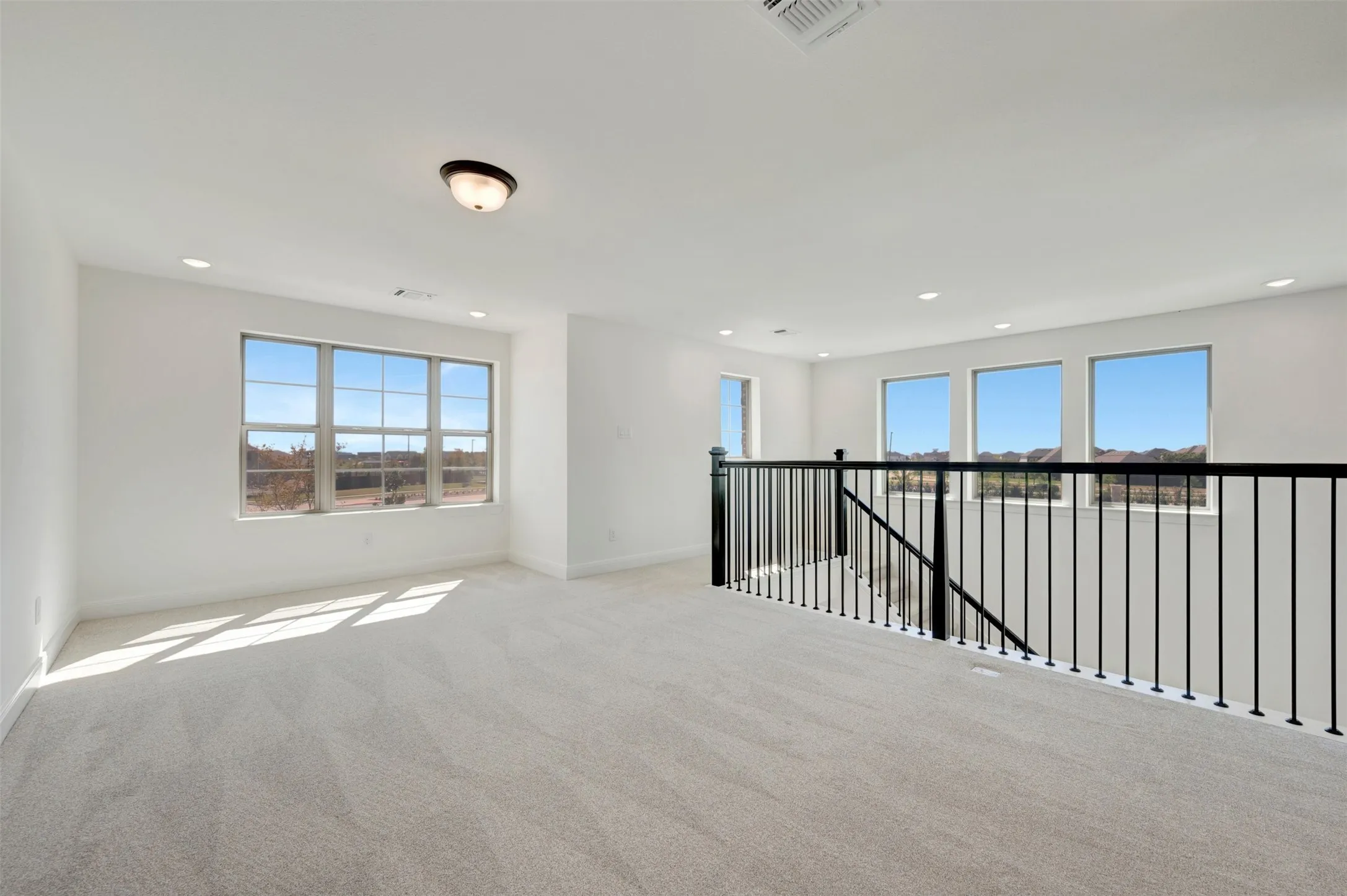 Expansive open loft area featuring light gray carpeting, multiple windows, recessed lighting, and a contemporary black metal railing