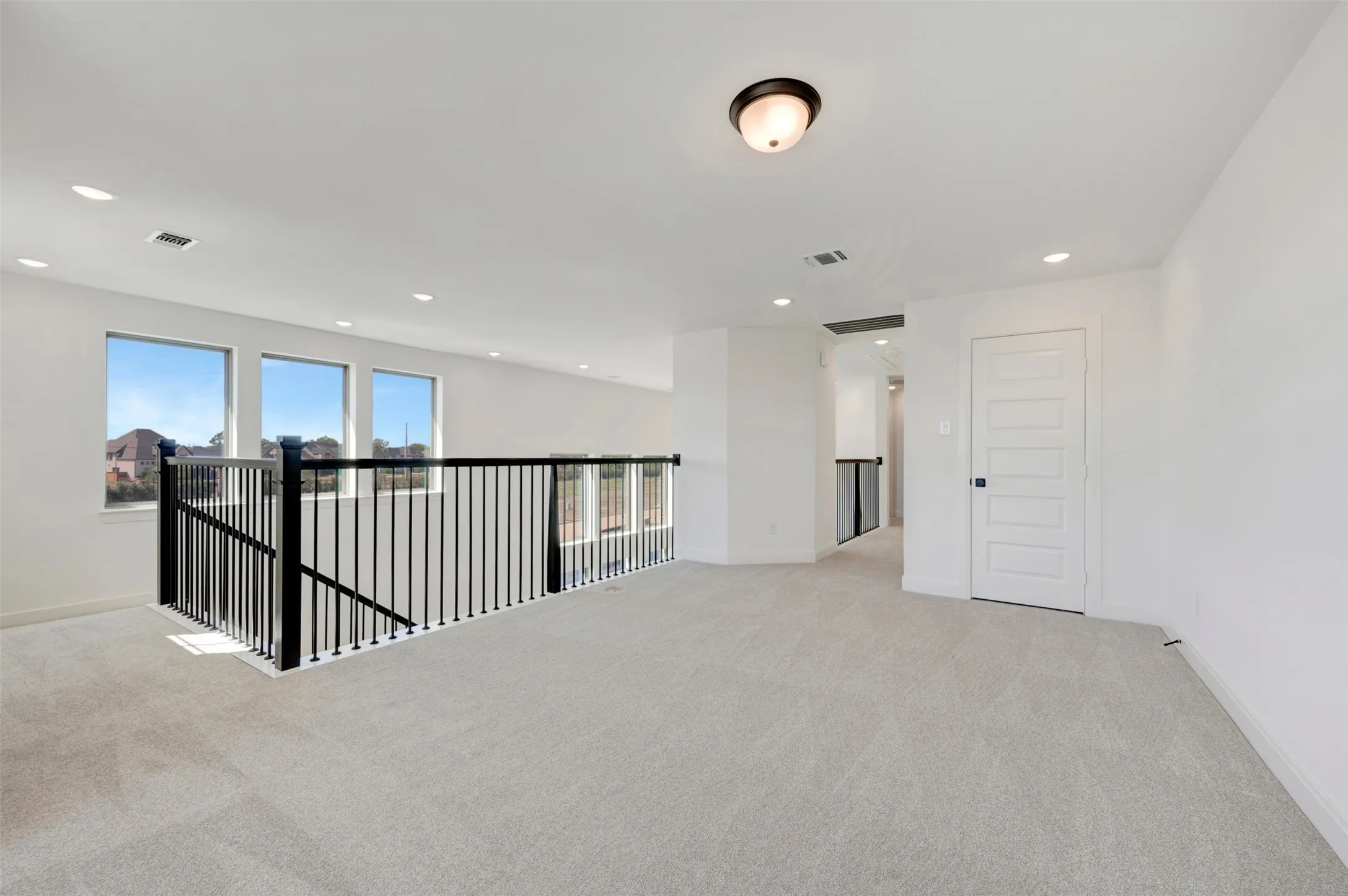 Spacious loft featuring light-toned carpeting, multiple windows, a black metal railing, a panel door, and recessed lighting