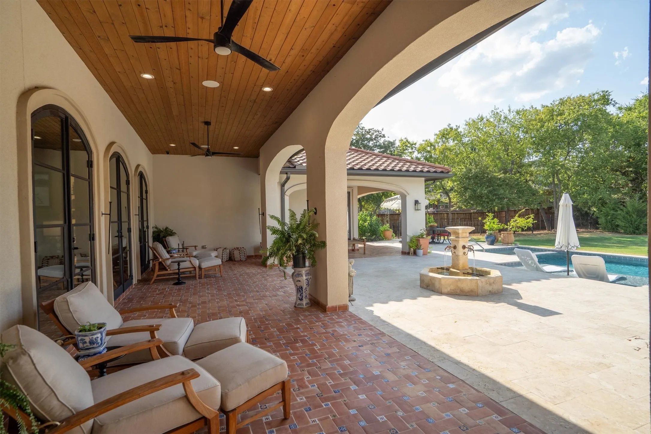 Expansive covered patio with wood ceiling, seating area, and tranquil fountain overlooking the pool.