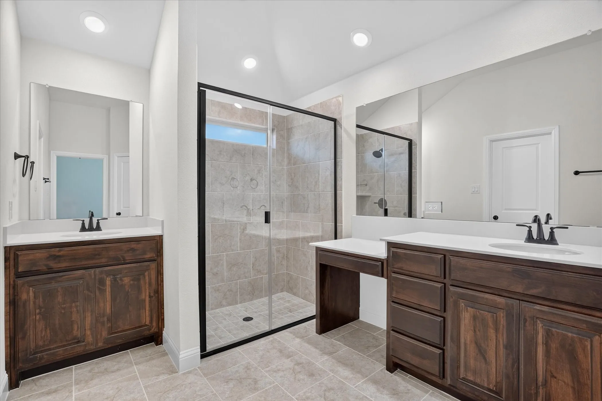 Spa-inspired primary bathroom featuring dual wood-finish vanities with vessel sinks and matte black hardware, frameless glass shower enclosure with black trim and stone tile surround, recessed ceiling lighting, and neutral tile flooring throughout