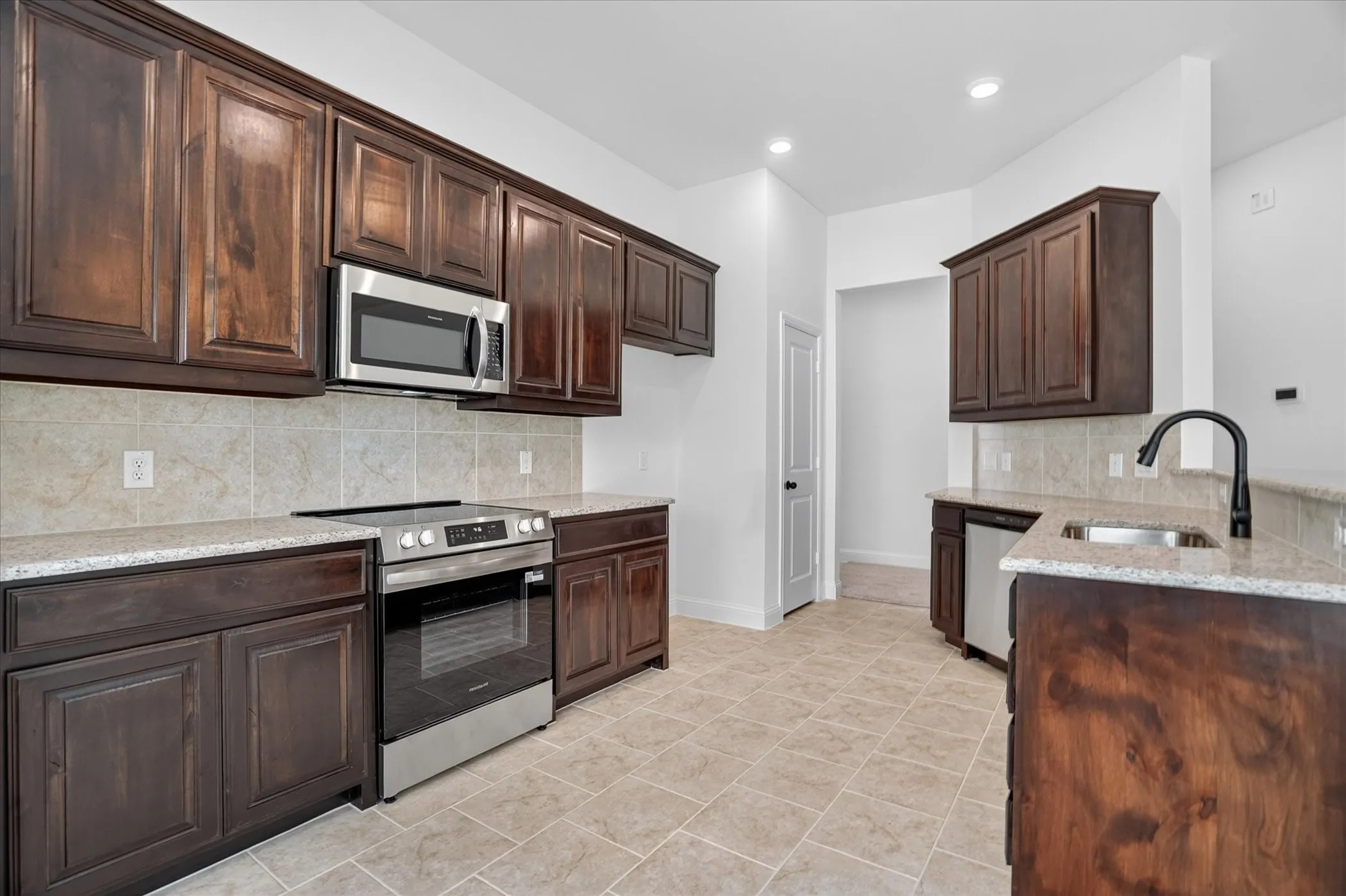 Kitchen featuring dark wood cabinetry, stainless steel appliances, light-toned tiled backsplash, speckled countertops, and tiled flooring