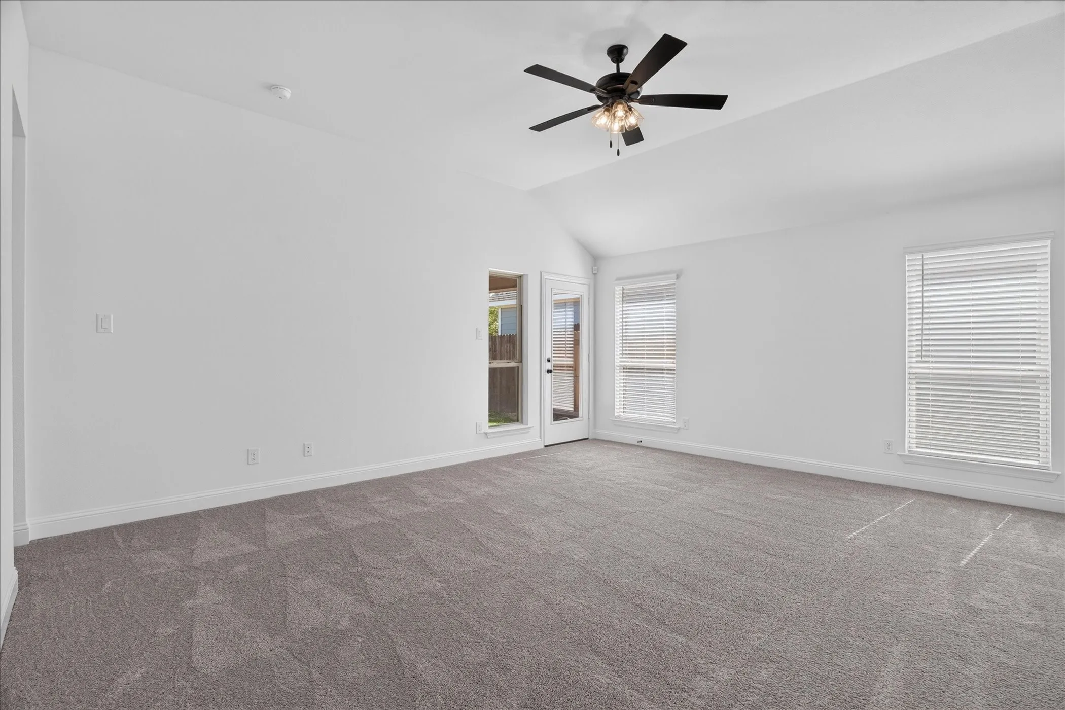 Spacious room featuring vaulted ceilings, light gray carpeting, multiple windows with blinds, and a ceiling fan