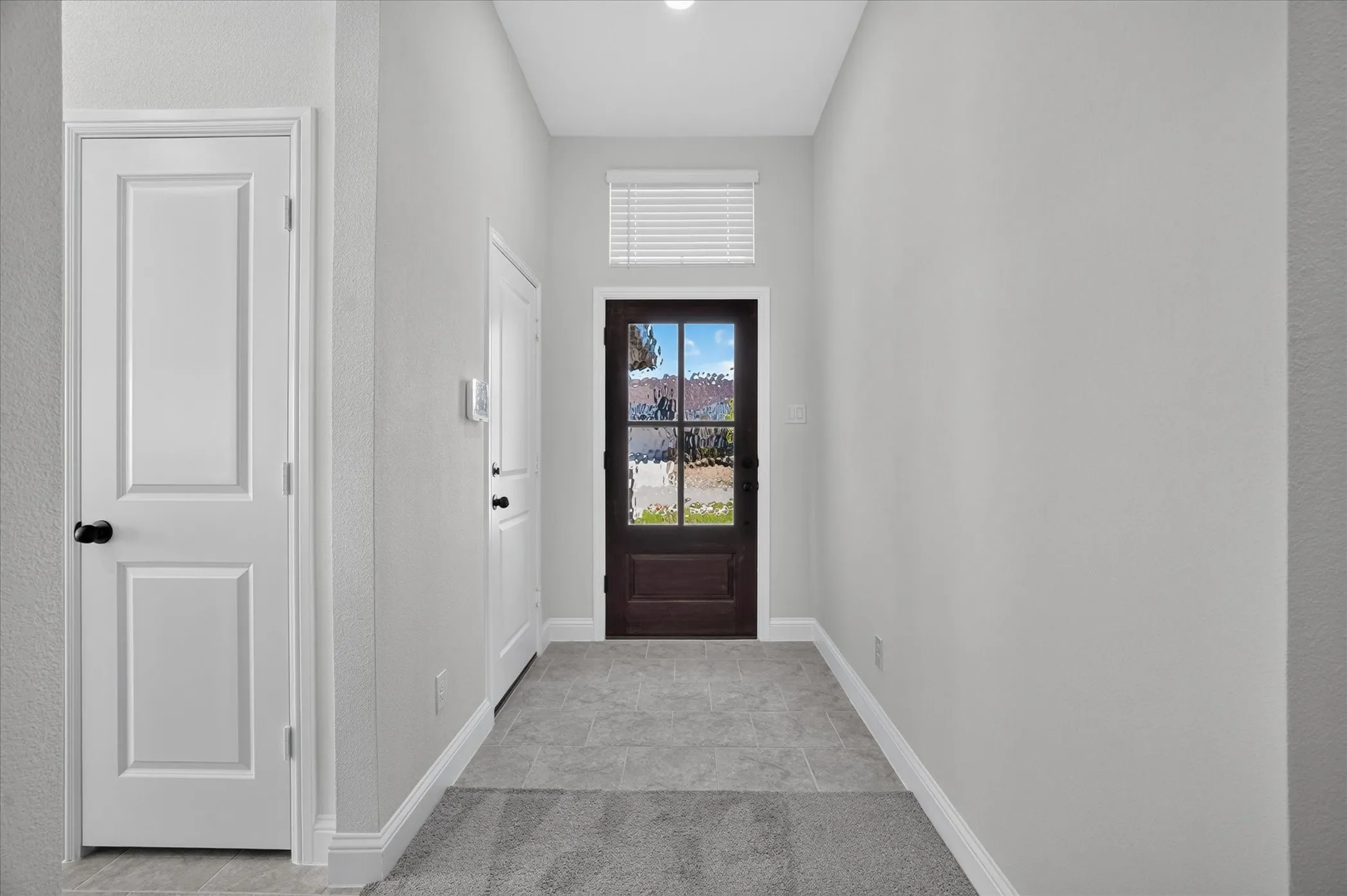 Recessed lighting in ceiling, white interior doors, dark wood-finish front door with glass panels, light gray tile flooring, and white baseboards