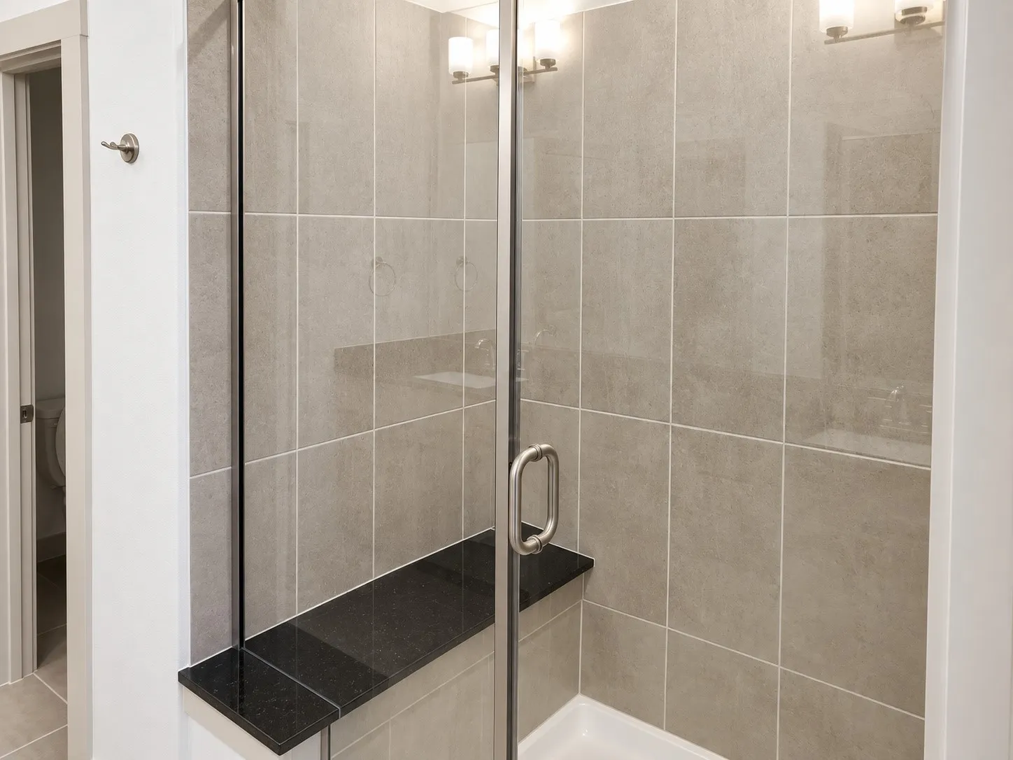Glass-enclosed shower with gray tiled walls, a dark stone shower bench, and a white shower pan