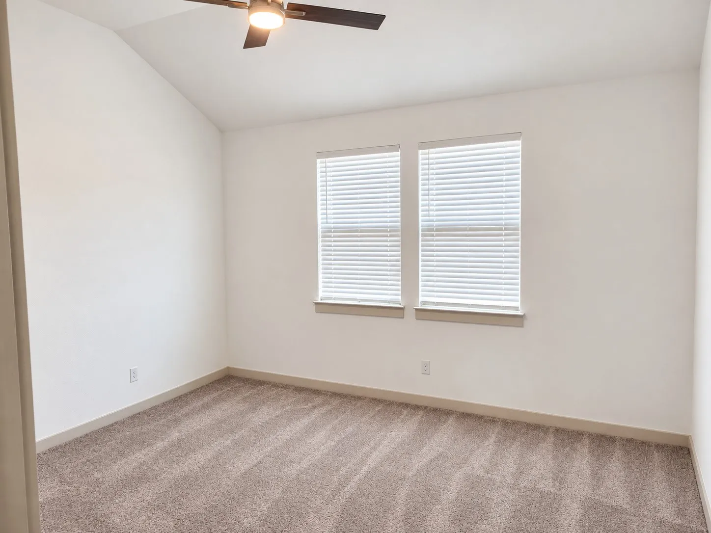 Vaulted ceiling, two windows with blinds, ceiling fan with integrated lighting, and neutral carpeting