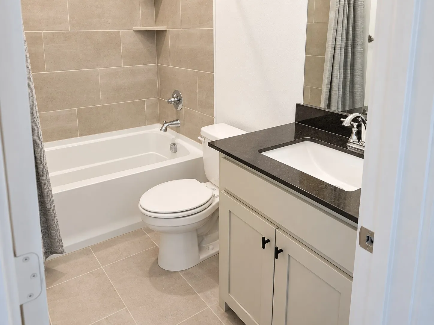 White acrylic tub with chrome fixtures, tiled shower surround with corner shelf, light-toned floor tiling, white vanity with two cabinet doors, and a dark countertop with an integrated sink