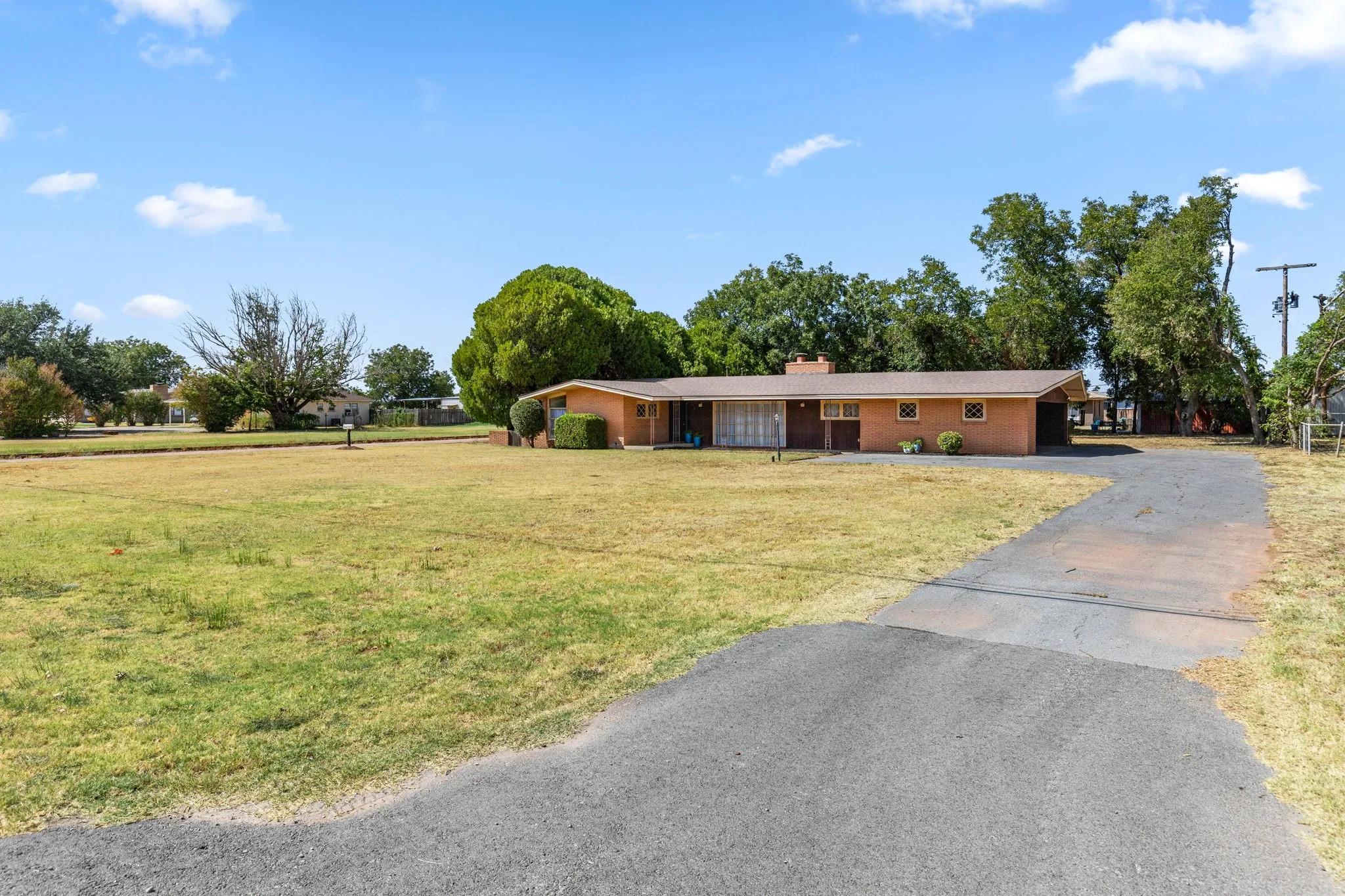 Brick exterior with a covered carport, mature trees, an expansive lawn, and a paved driveway
