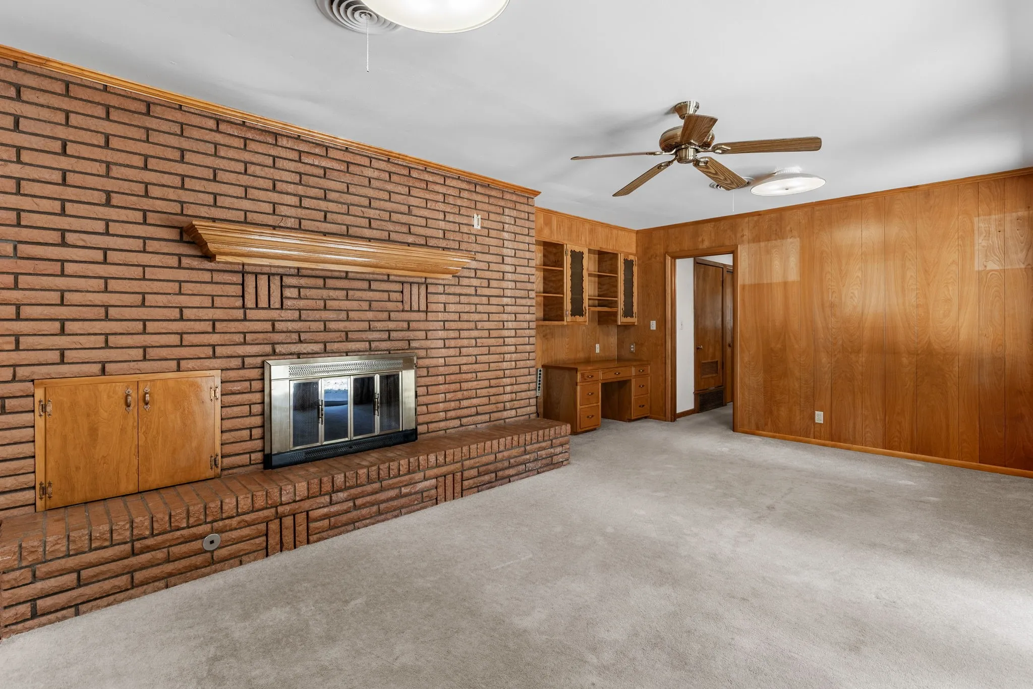 Brick-clad fireplace with mantel and hearth, built-in desk with shelving, wood paneling, and ceiling fan