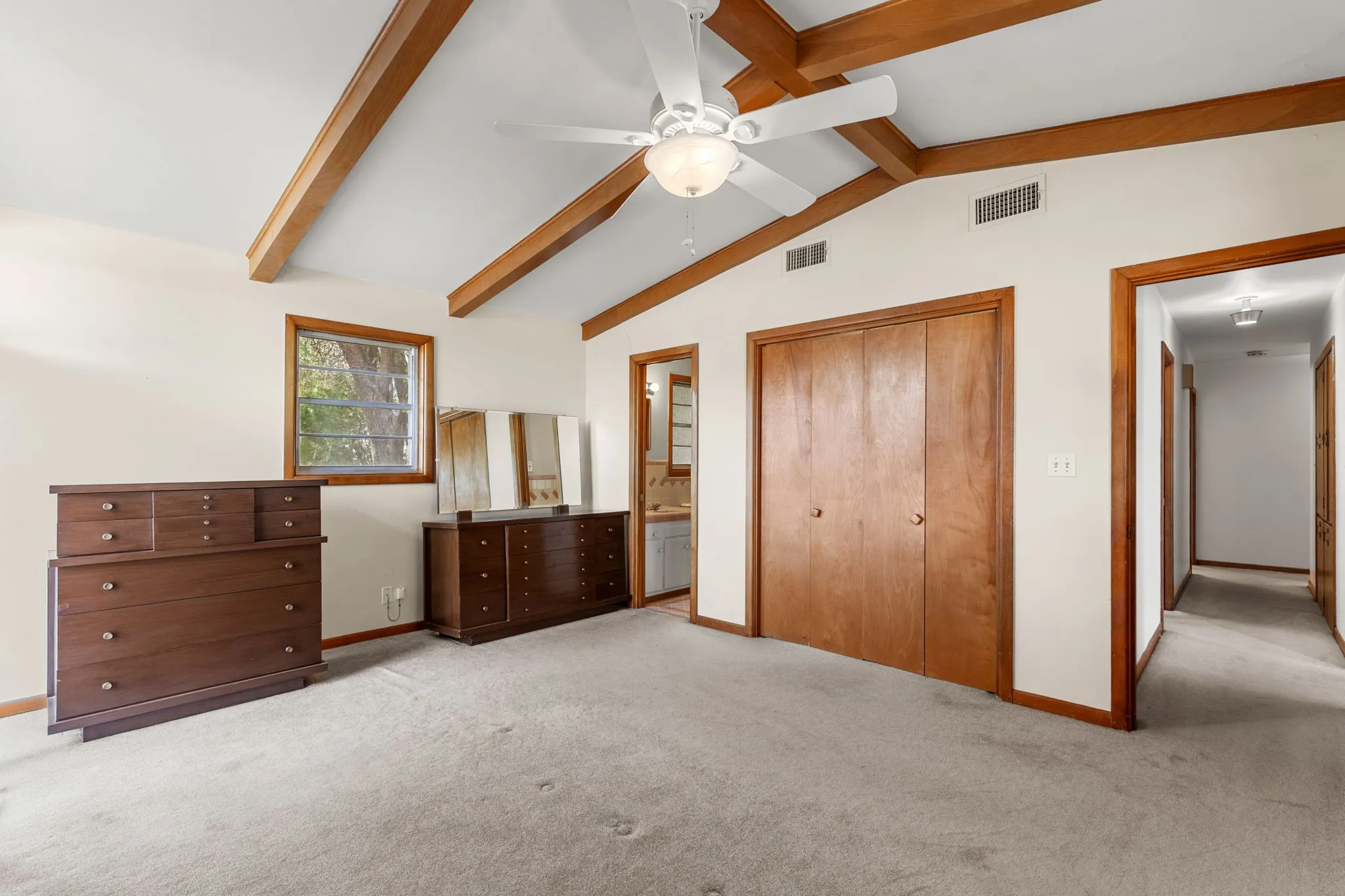 Vaulted ceiling with exposed wood beams, ceiling fan with integrated lighting, wood-finish bi-fold closet doors, and wood-trimmed window