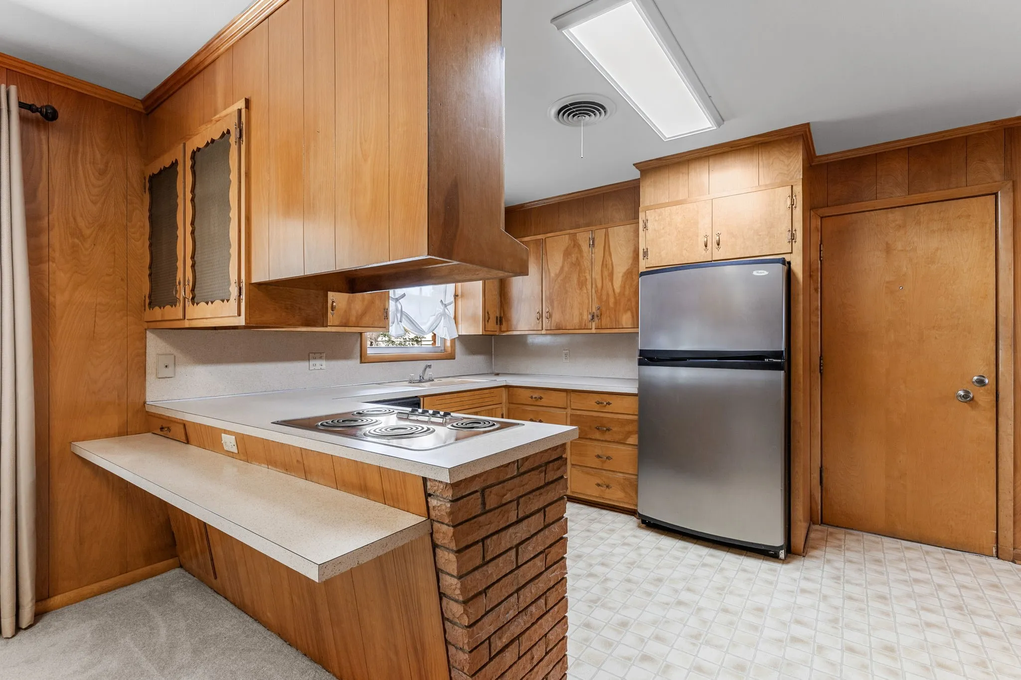Wood-paneled kitchen featuring an integrated cooktop, brick-finish island base, stainless steel refrigerator, and ample cabinetry