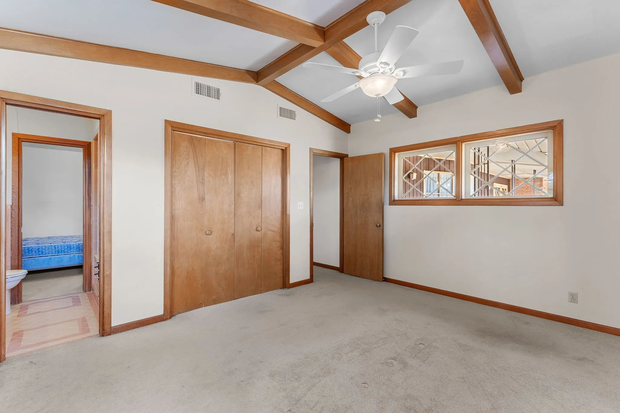 Vaulted ceiling with exposed wood beams, white ceiling fan with integrated lighting, wood-framed windows, and wood-paneled closet doors