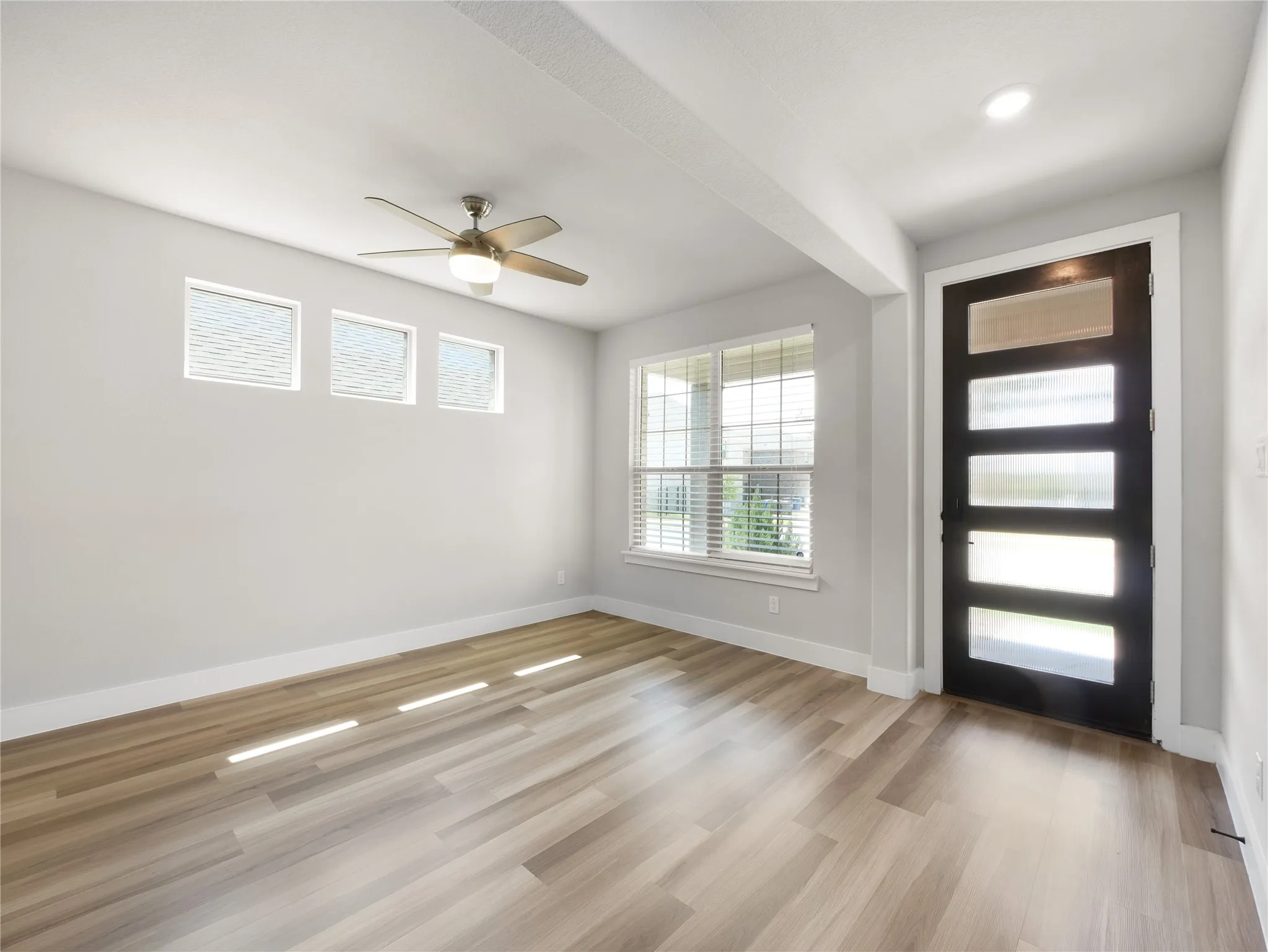 Wood-finish flooring, a contemporary black entry door with frosted glass panels, a ceiling fan with integrated lighting, and multiple windows with blinds