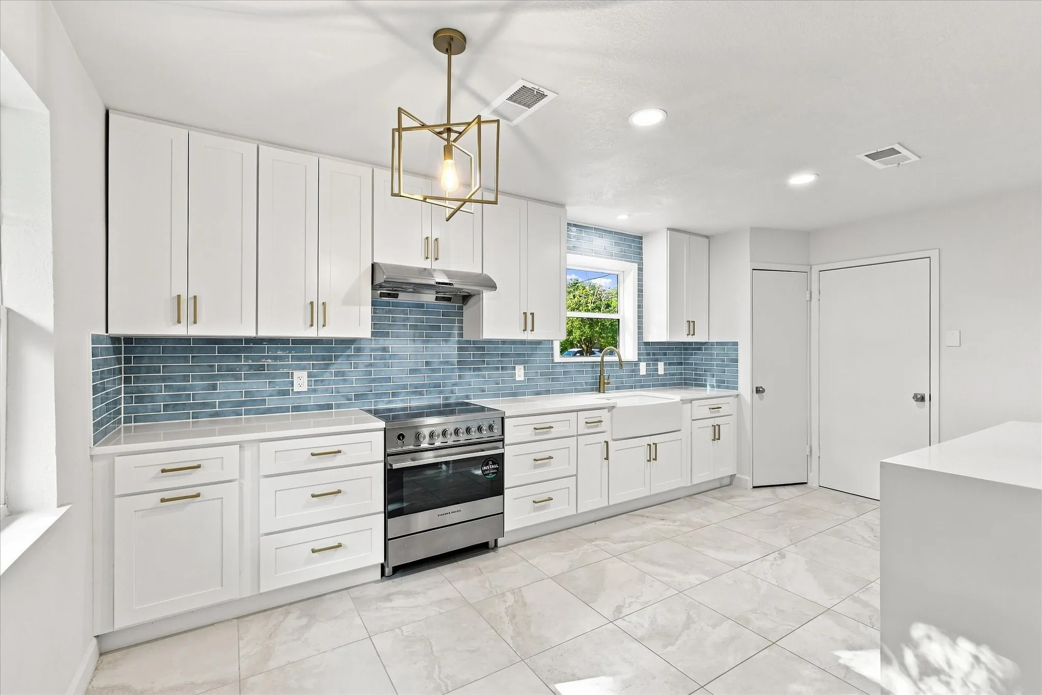 Bright kitchen featuring white shaker cabinets, a full-height blue tile backsplash, stainless steel range, farmhouse sink, and a modern geometric pendant light with pantry