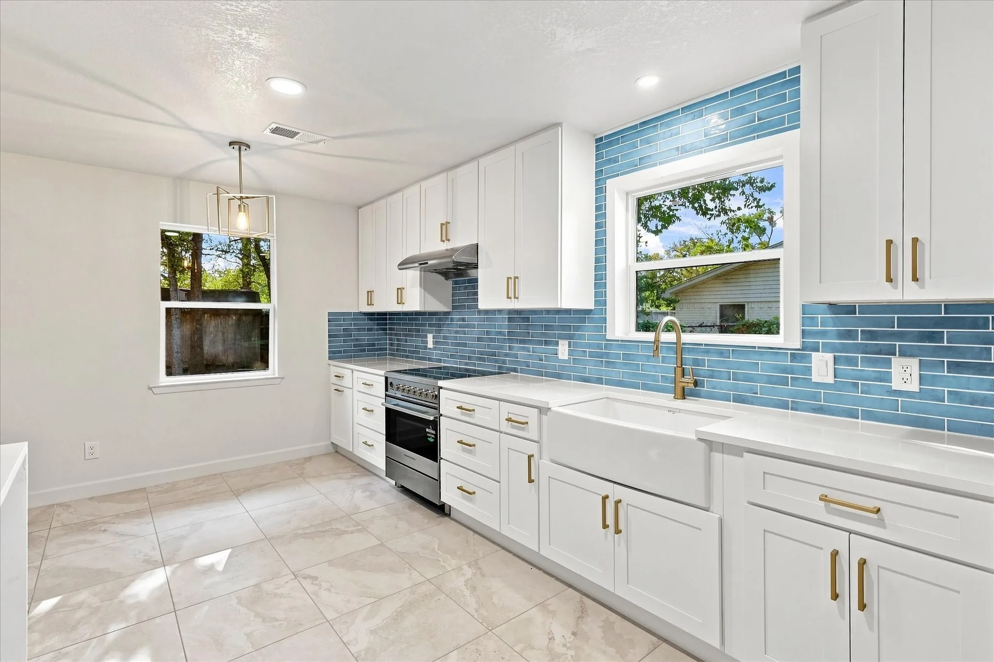 Kitchen featuring white cabinetry with gold-toned hardware, full blue tile backsplash, stainless steel range with overhead hood, and a farmhouse sink