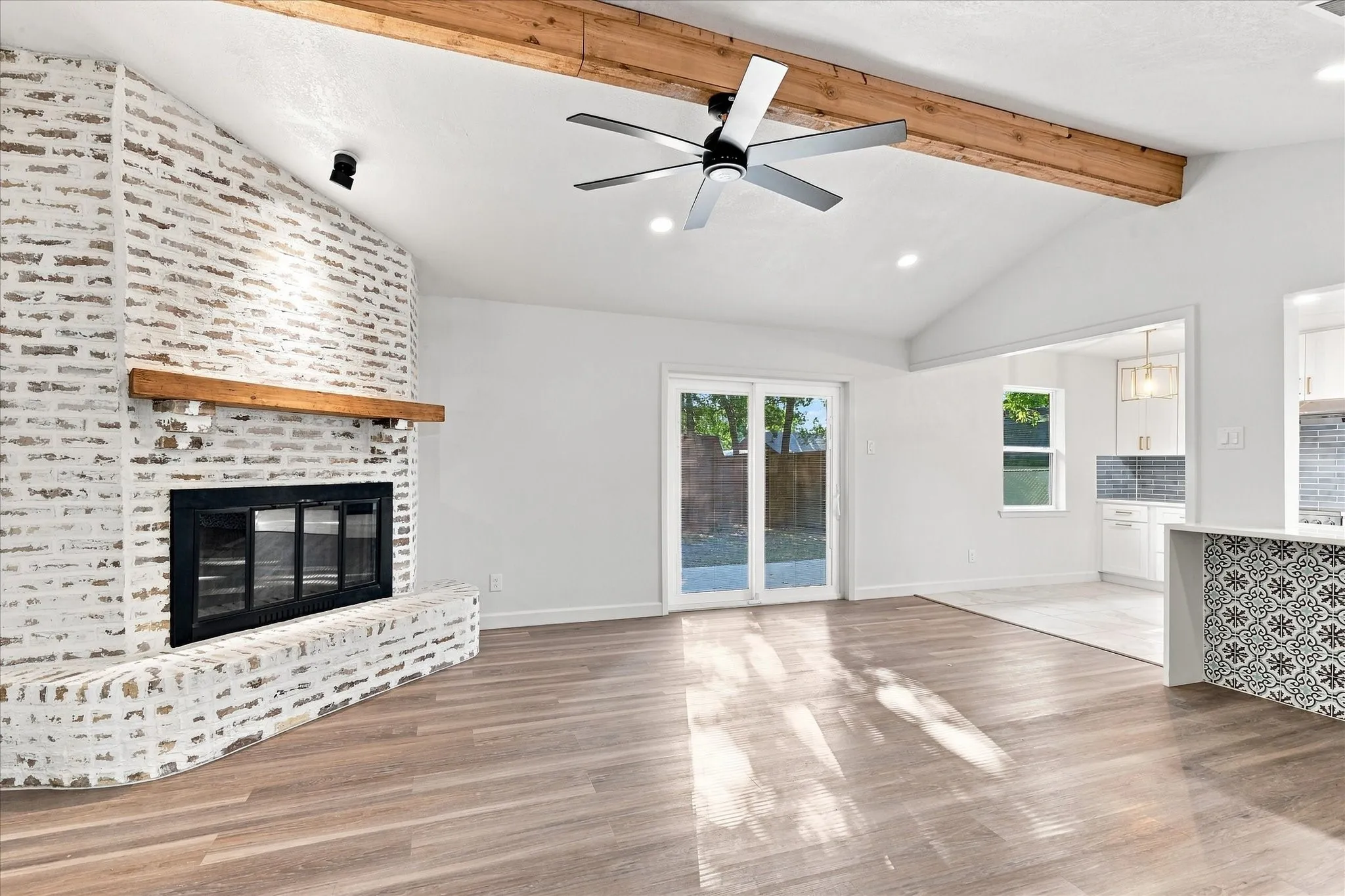 Vaulted ceiling with exposed wood beams, wood-finish flooring, brick fireplace with wood mantel, ceiling fan, and sliding glass doors