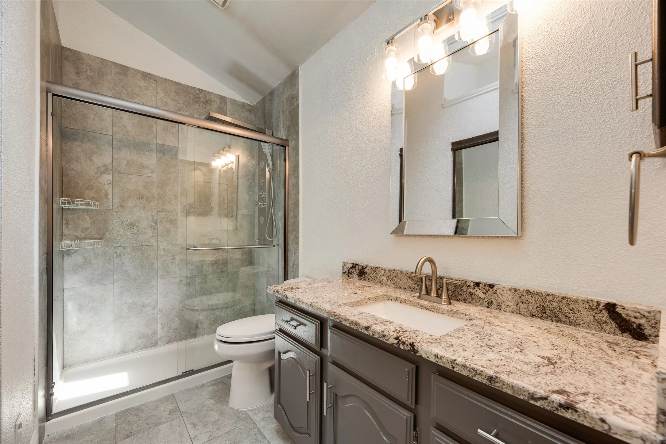 Walk-in shower with tiled surround and glass enclosure, white toilet, vanity with undermount sink, granite countertop, and brushed nickel fixtures