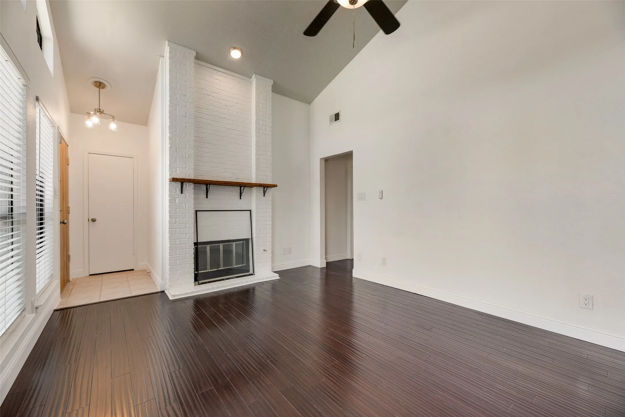 Vaulted ceiling with an installed ceiling fan, wood-finish flooring, a white brick fireplace with a wood mantle, and an entry with tile flooring