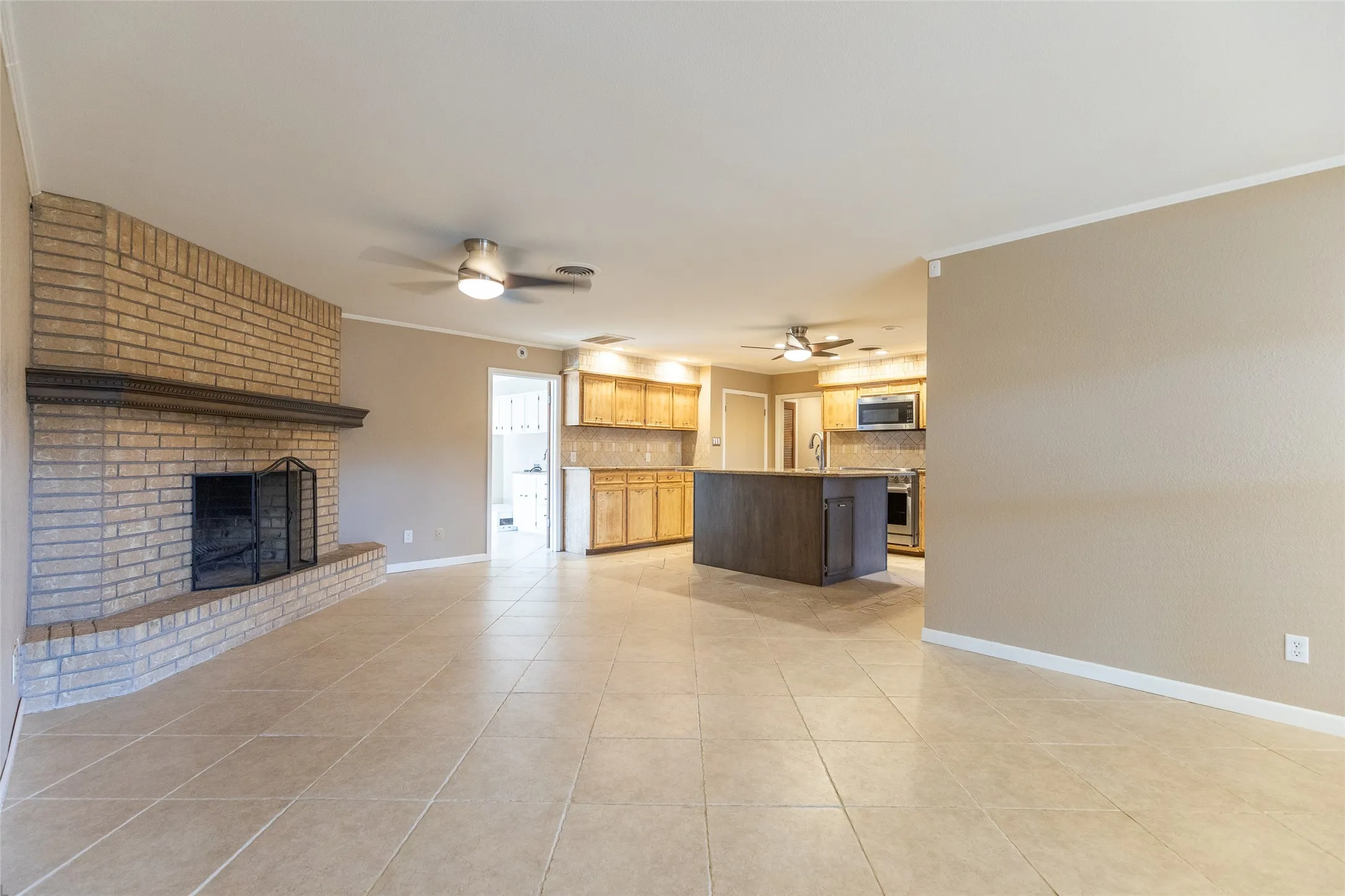 Tile flooring, brick fireplace with hearth, ceiling fans with integrated lighting, kitchen island with built-in cabinetry, and wood-finish kitchen cabinetry
