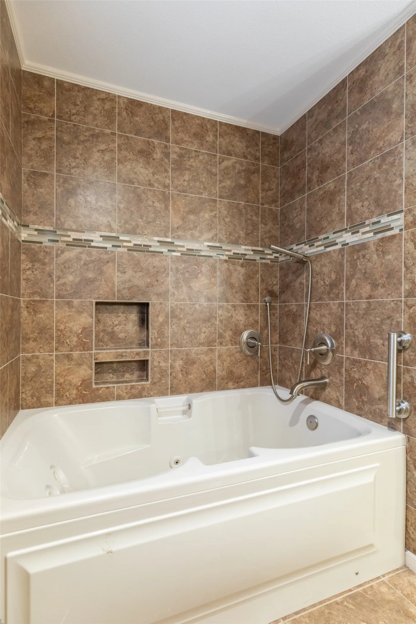 Jetted soaking tub with a tiled surround, decorative accent tile band, built-in shower niche, and grab bar