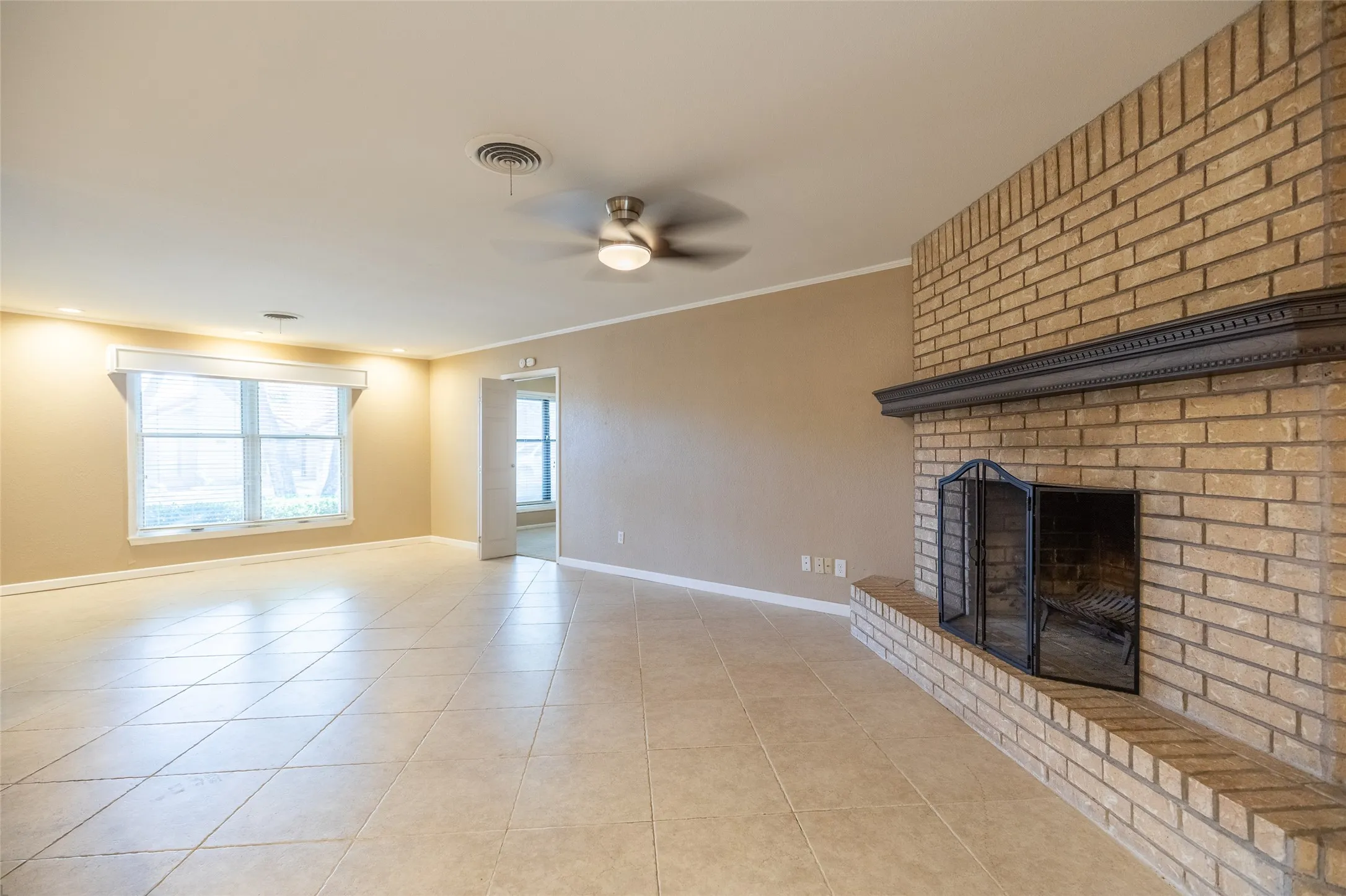 Spacious living area featuring a prominent brick fireplace with a dark mantel, large window, ceiling fan, and tile flooring