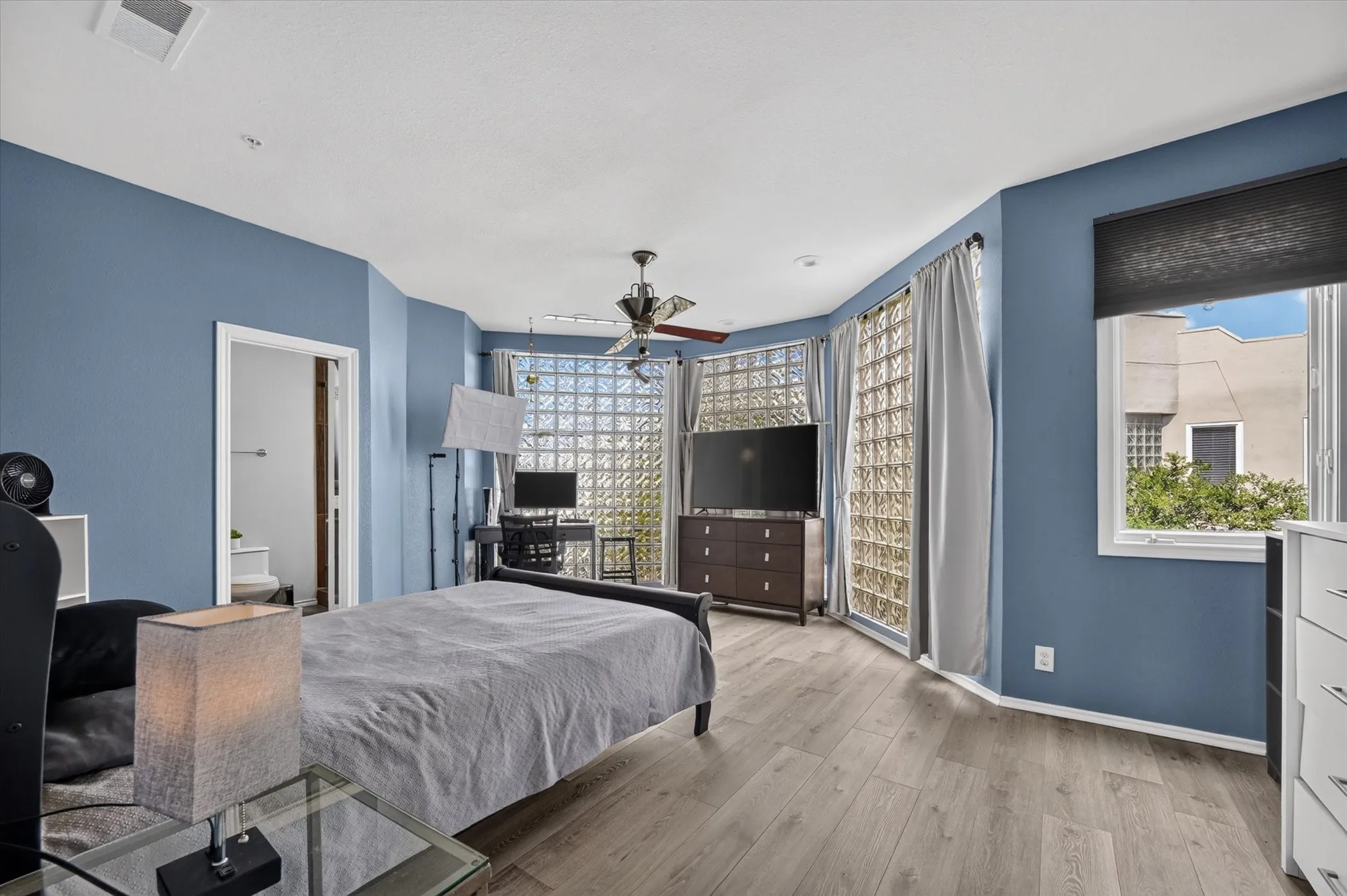 Wood-finish flooring, ceiling fan, and multiple windows including a large window with roller shade and floor-to-ceiling glass block panels