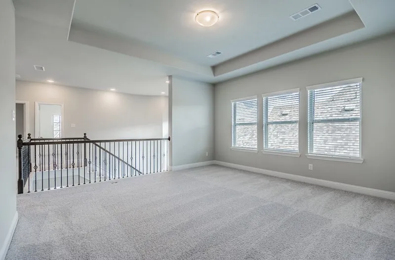 Spacious room with a tray ceiling and recessed lighting, plush carpet flooring, three windows with blinds, and an iron balustrade