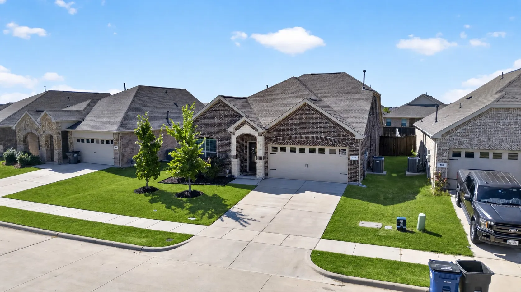 Brick and stone facade, attached garage with panel doors, concrete driveway, and manicured front lawn with mature trees