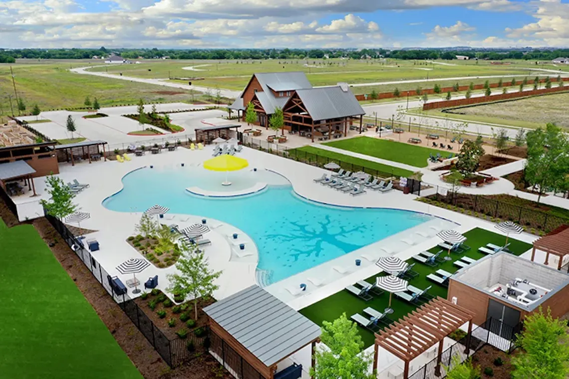 Expansive outdoor pool with integrated shallow area, adjacent amenity building with covered patio, multiple lounging areas, and a large manicured lawn