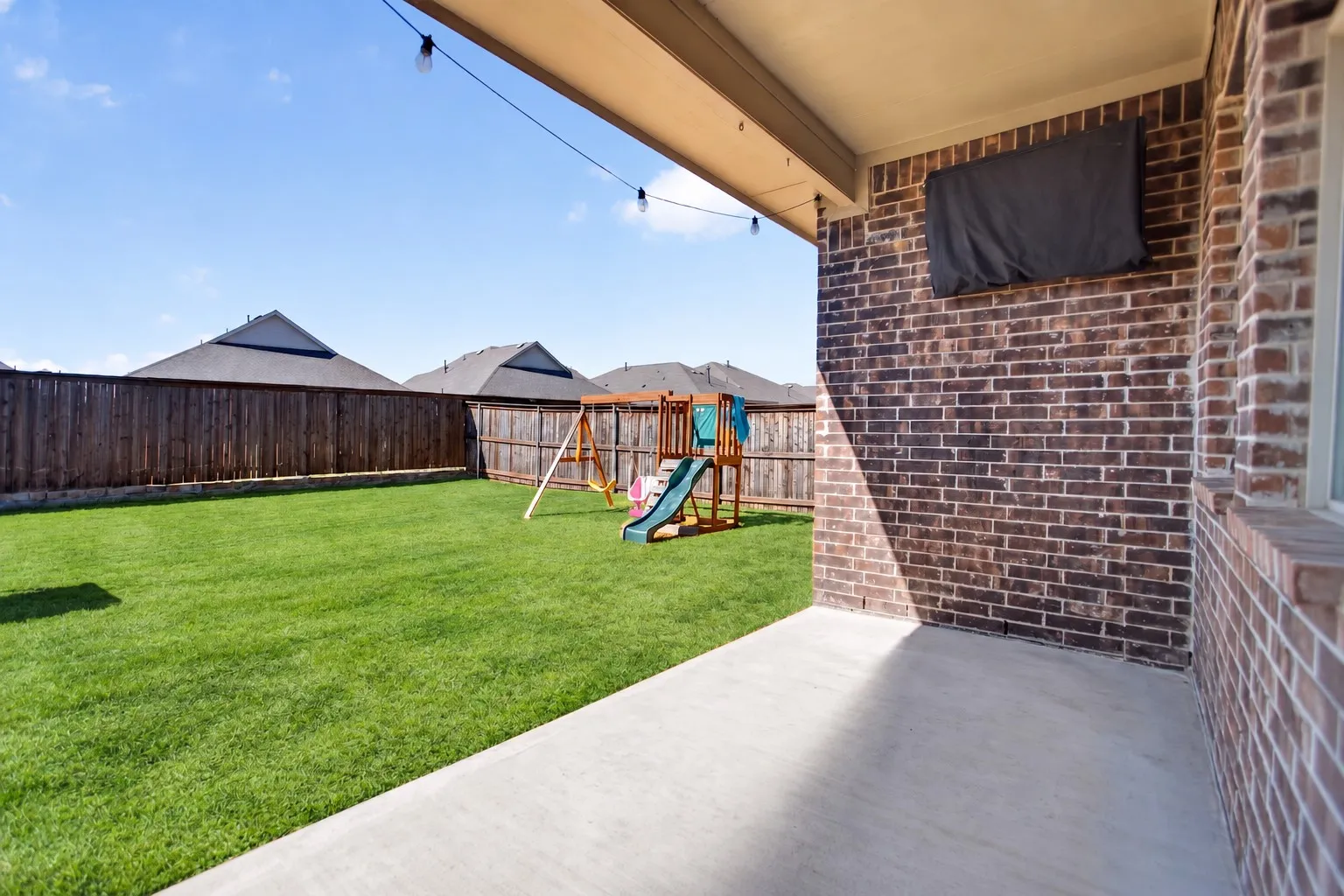 Expansive backyard featuring a manicured lawn, wood privacy fencing, and a covered concrete patio with brick wall
