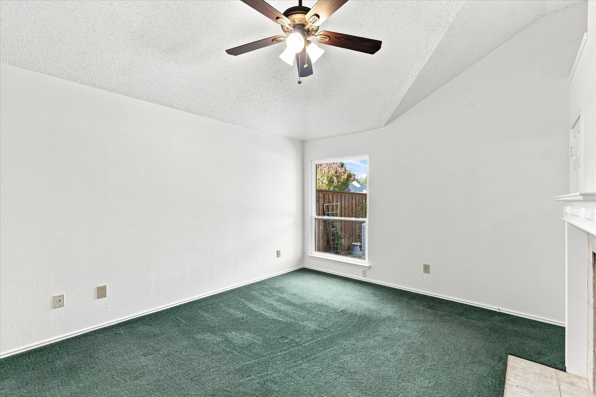 Vaulted ceiling with a ceiling fan, white interior walls, window with a white frame, green carpeting, and a fireplace with a white mantel