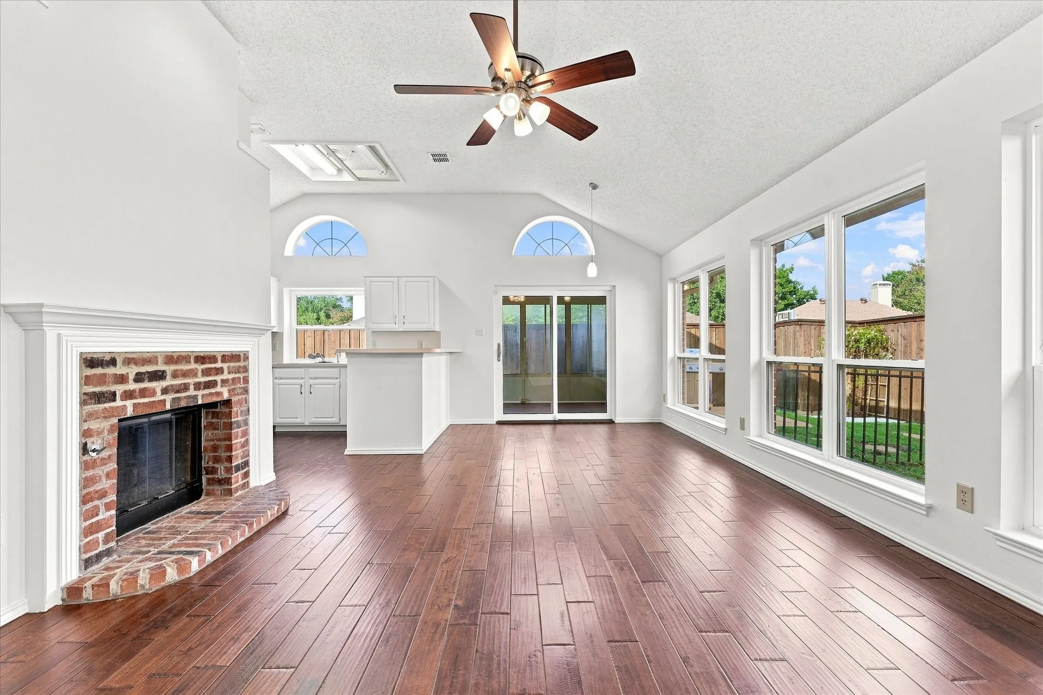 Living room to kitchen & dining area featuring engineered hardwood flooring, a brick fireplace, vaulted ceiling with a ceiling fan, and multiple windows